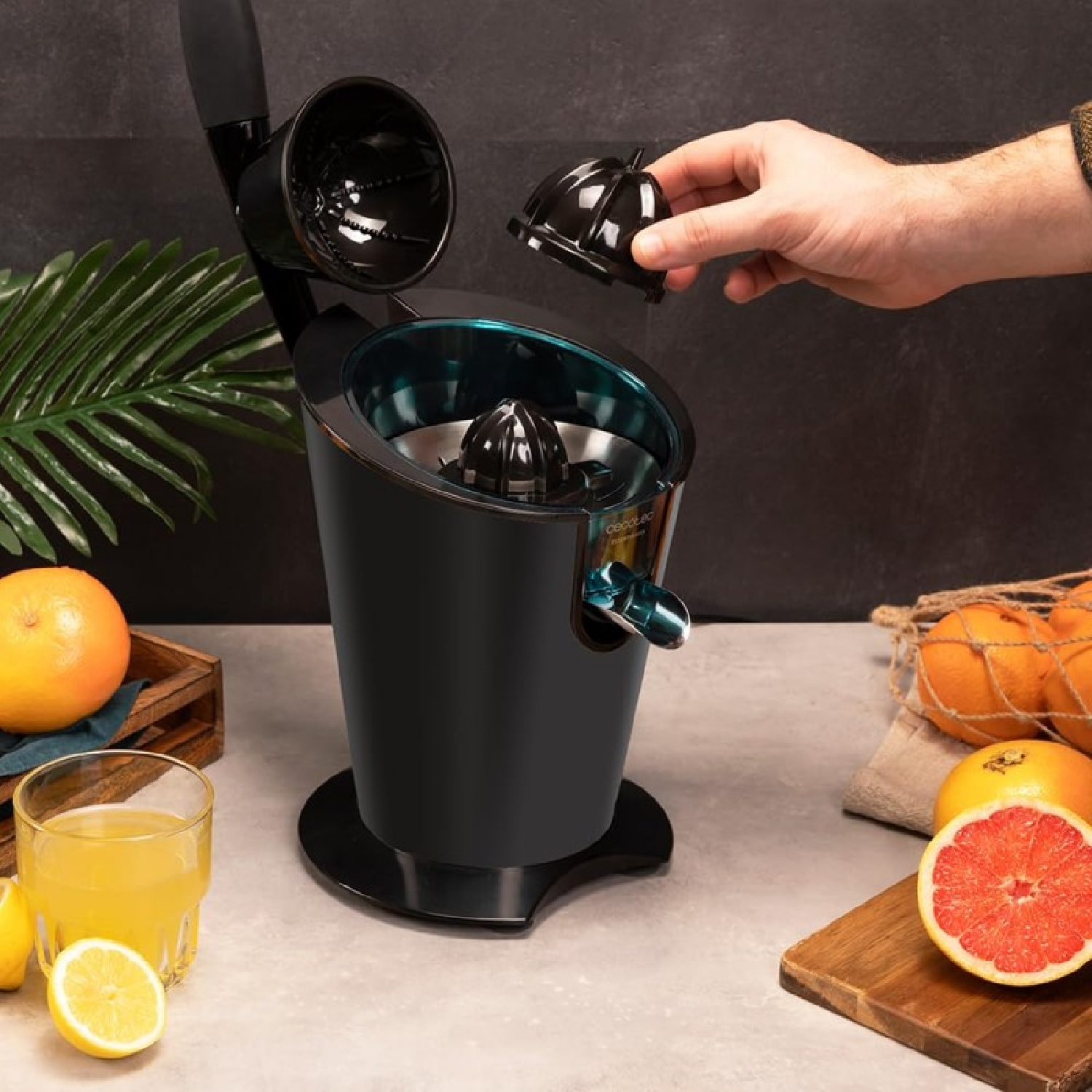 Una mano sostiene una parte de un exprimidor eléctrico negro junto a naranjas y un vaso de zumo en una mesa.