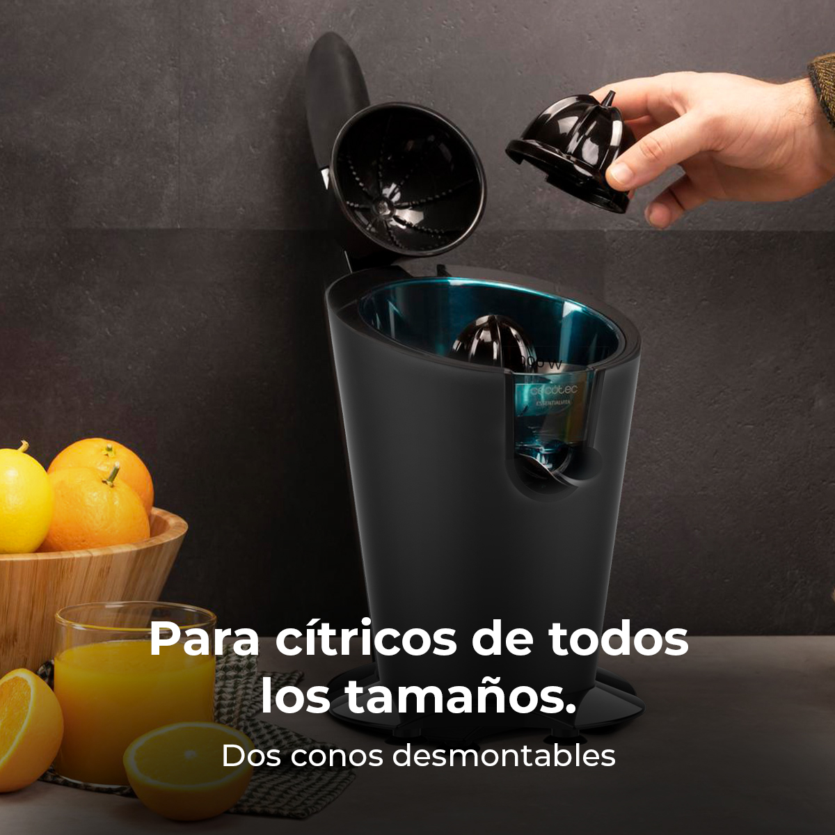 Una mano coloca un cono exprimidor negro en un exprimidor junto a un bol de naranjas y un vaso de zumo.