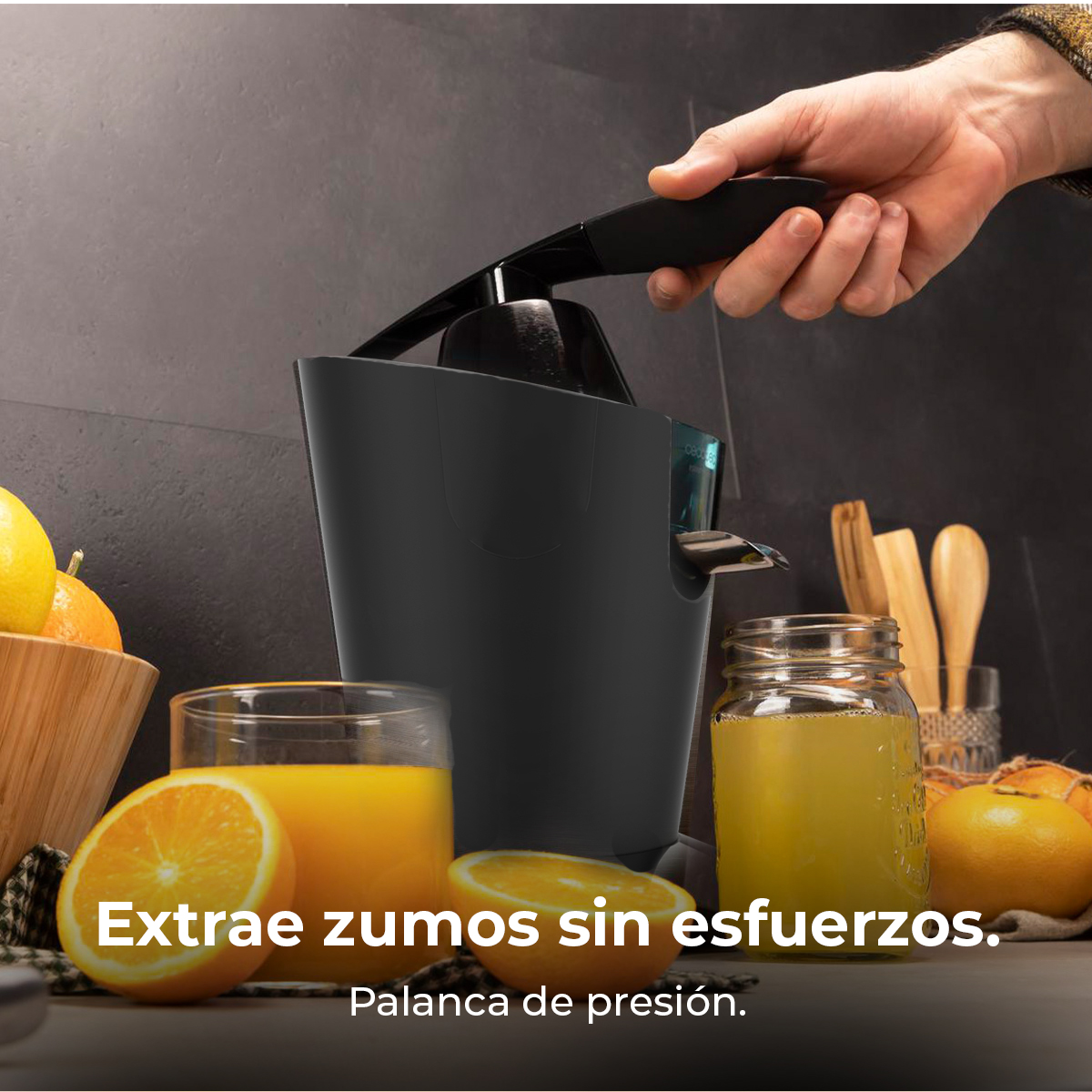 Mano presionando la palanca de un exprimidor negro junto a vasos de zumo de naranja y naranjas.