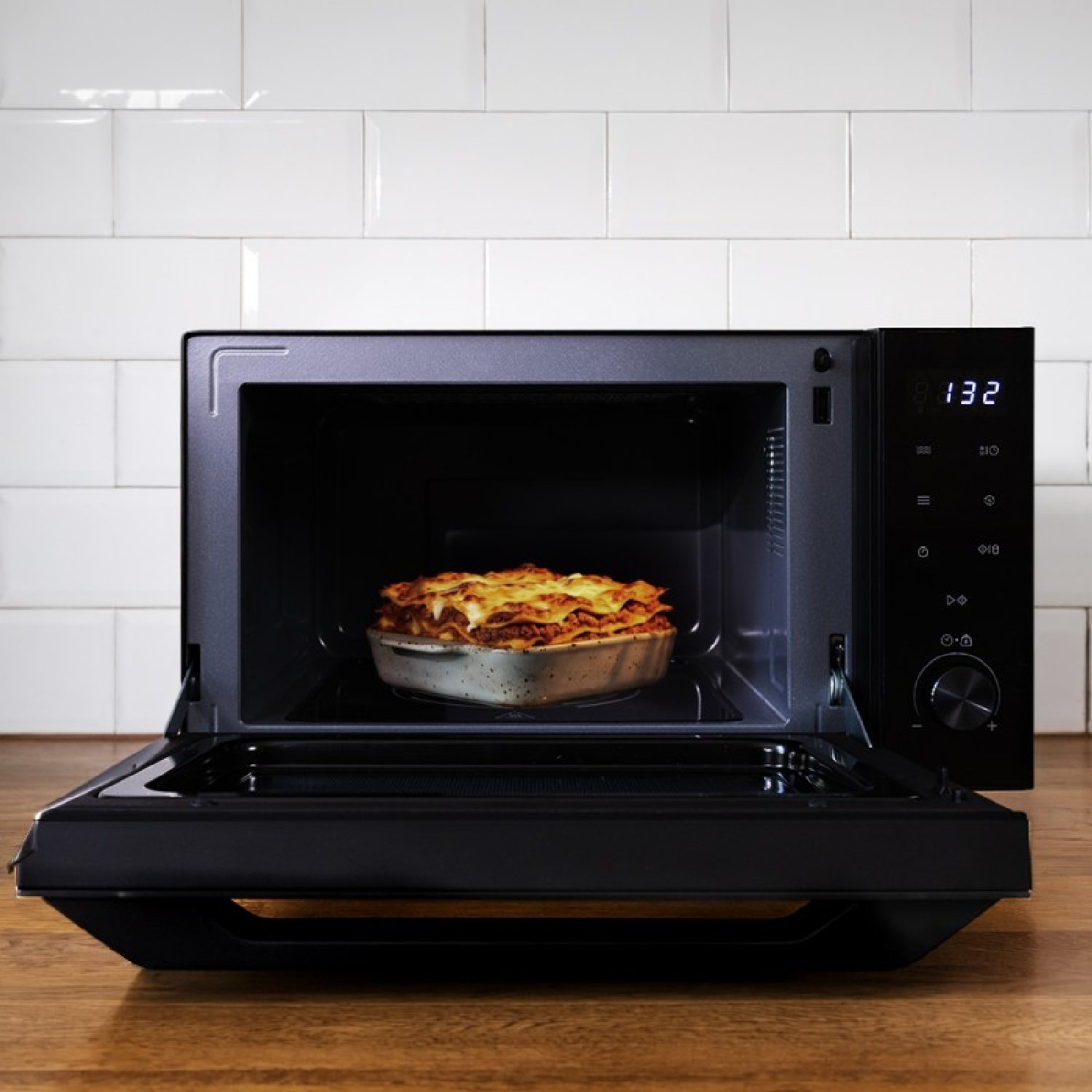 Una lasaña dentro de un horno microondas negro abierto con azulejos blancos.