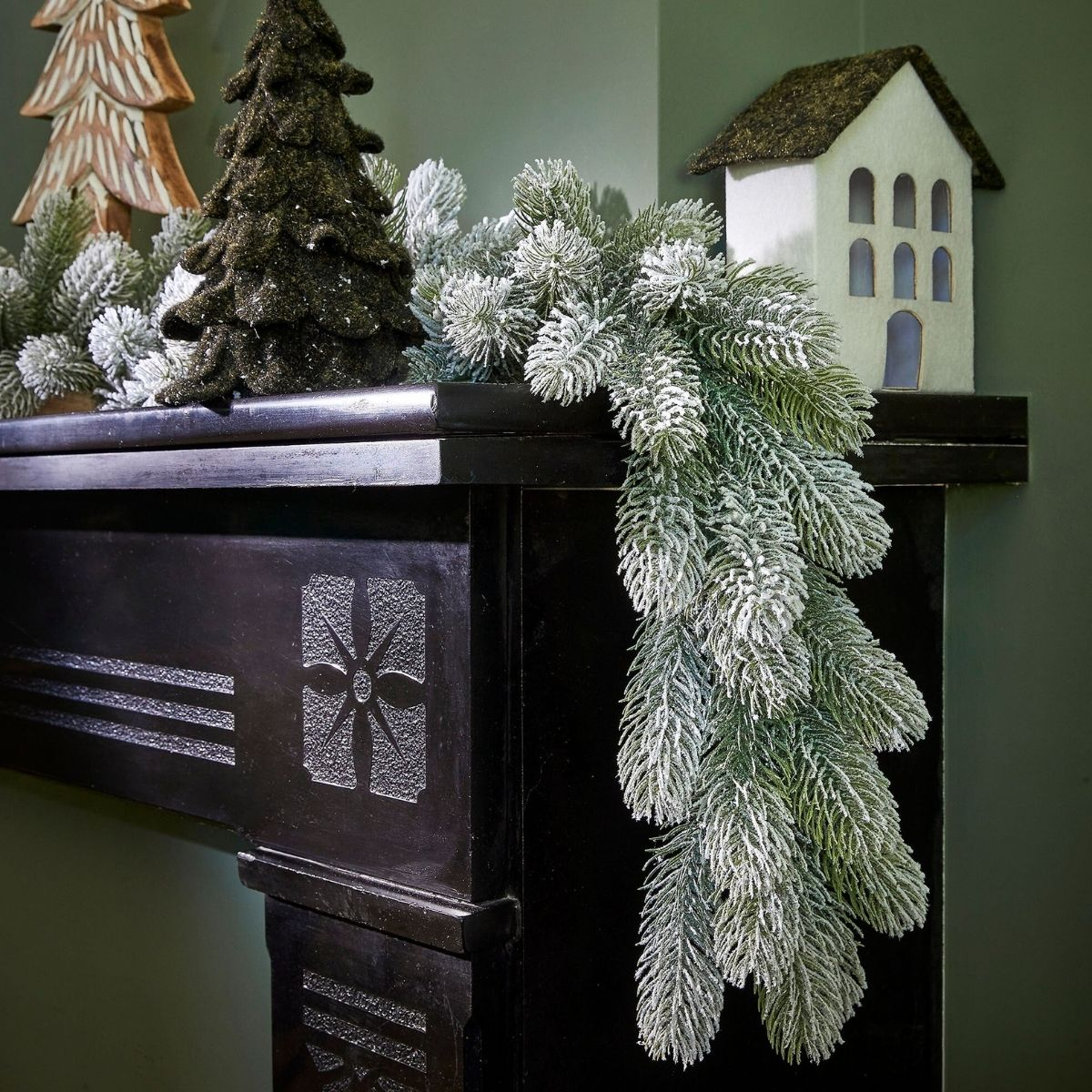 Une guirlande de pin enneigée, des arbres sombres et un ornement de maison blanche décorent un manteau de cheminée noir.