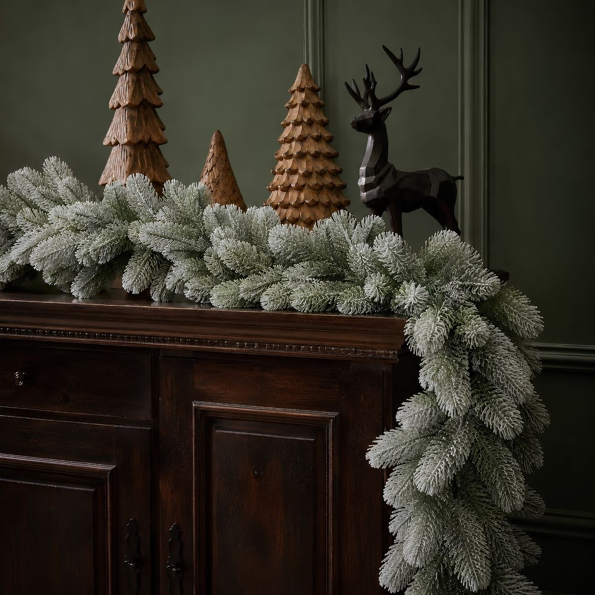 Un buffet en bois sombre avec guirlande de sapin enneigée, sapins en bois et renne sombre sur fond vert.