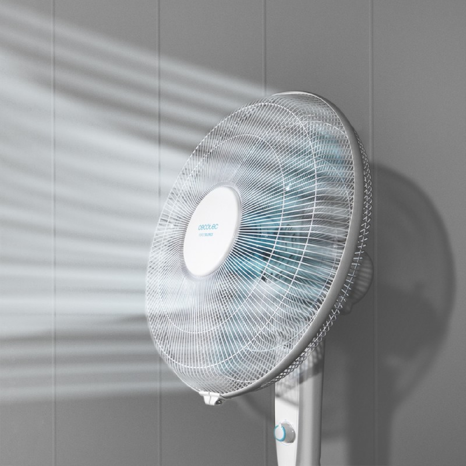Un ventilador de pie blanco sopla viento contra una pared gris con rayos de luz.