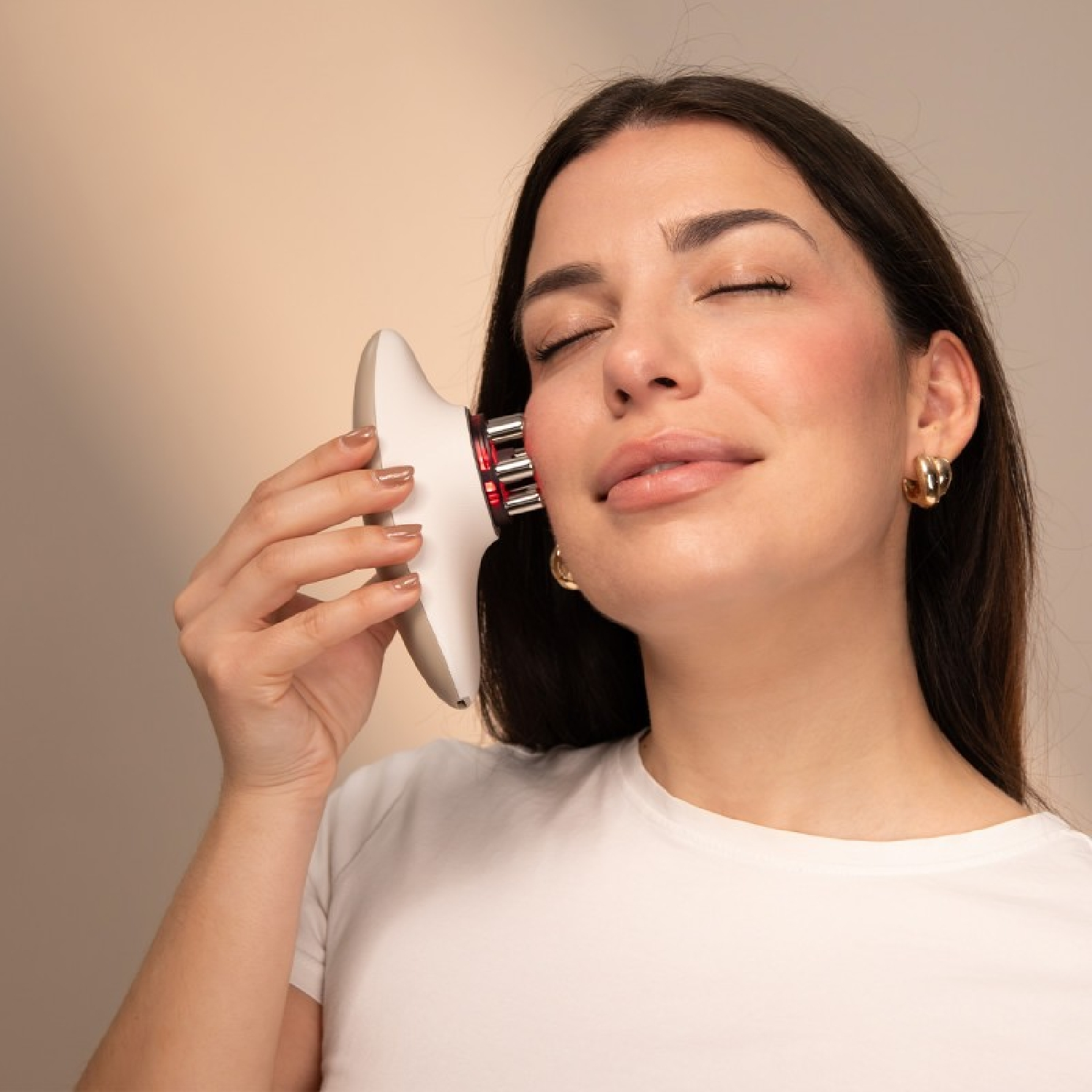 Una mujer sostiene un dispositivo facial blanco con accesorios de metal en la mejilla.