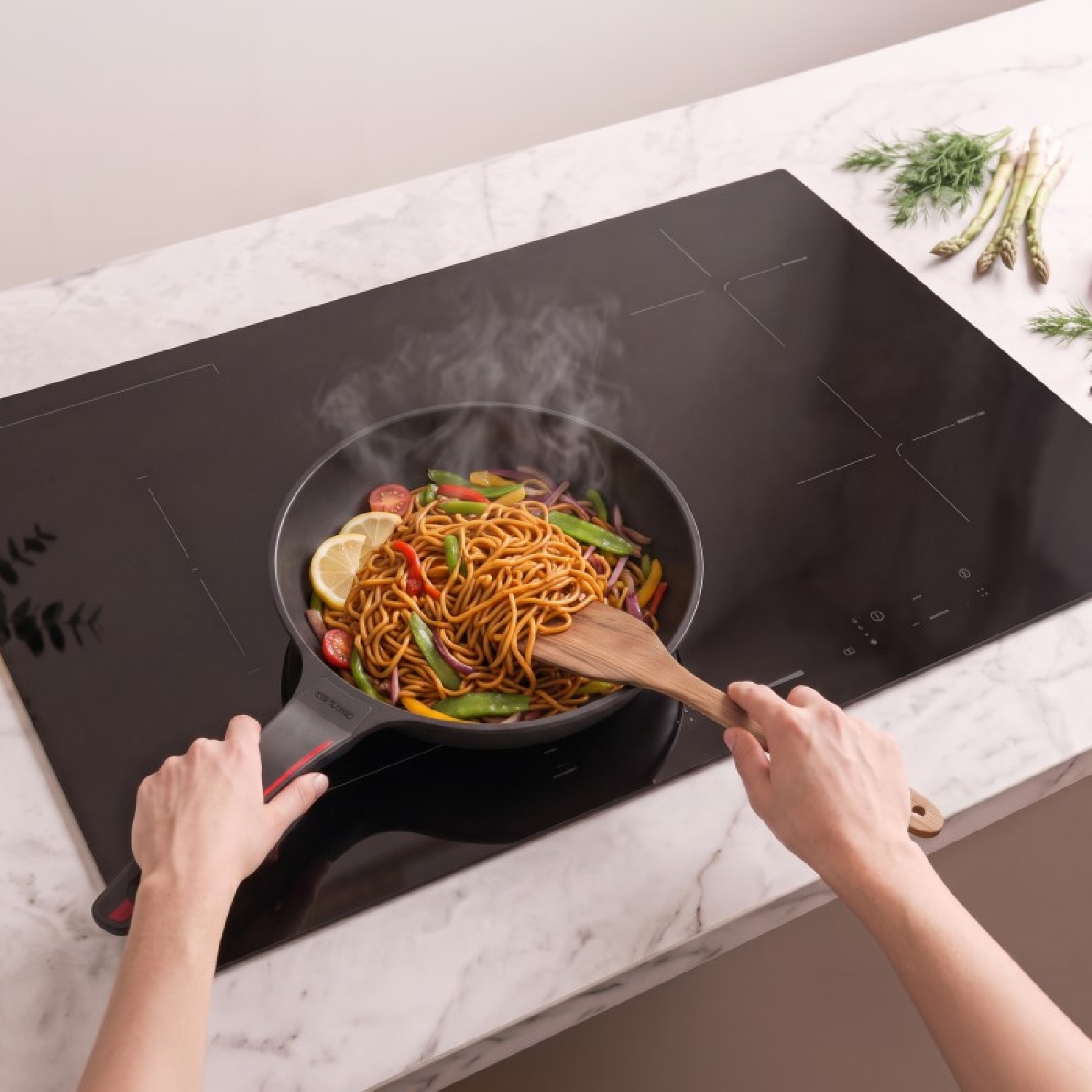 Persona revuelve fideos humeantes con verduras en una sartén sobre una placa de vitrocerámica.