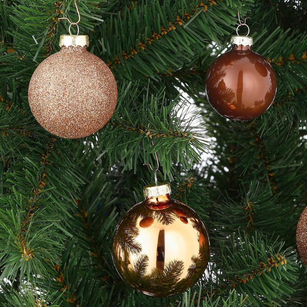 Three Christmas baubles hanging on a green artificial pine tree branch. One is sparkly, the other two are glossy.
