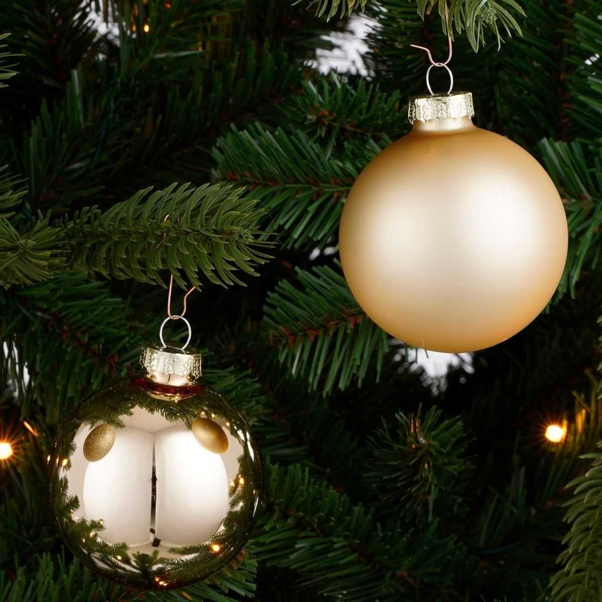Two gold Christmas baubles hang on green branches of a Christmas tree with small lights.