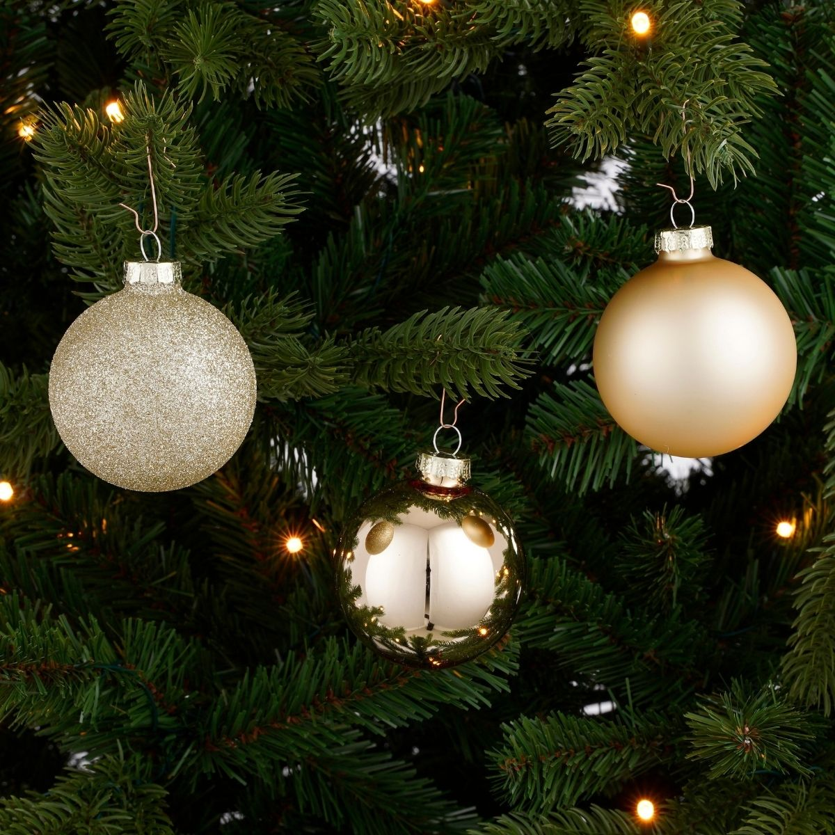 Three gold Christmas ornaments hang on a green fir tree with glowing lights.