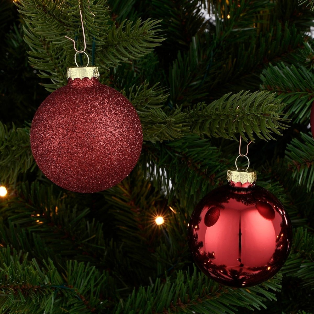 Two red Christmas ornaments hang on a green Christmas tree with lights.