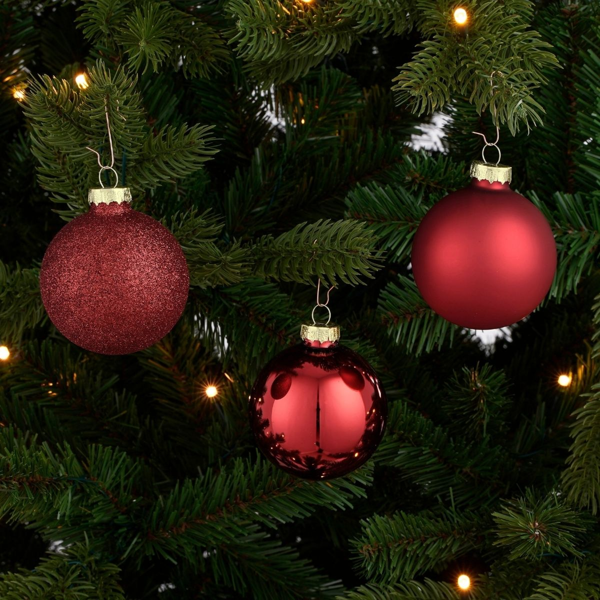 Three red Christmas ornaments hang from the branches of a fir tree, surrounded by small lights.