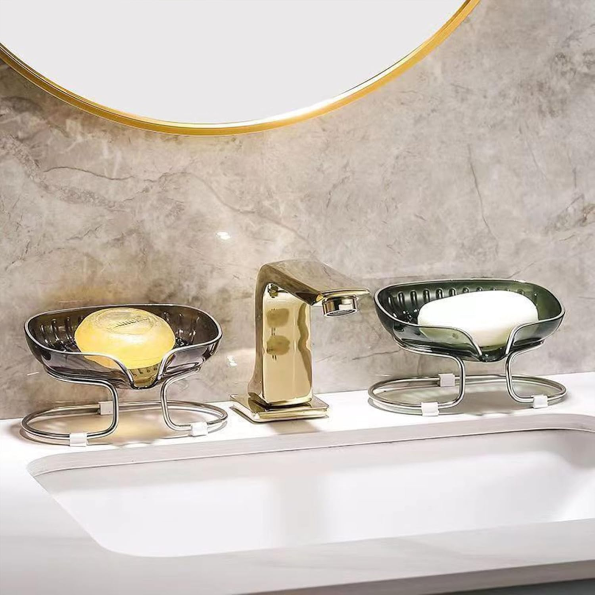 Zwei graue Seifenschalen mit Seife auf einem Waschbecken neben einem goldenen Wasserhahn an einer Marmorwand.