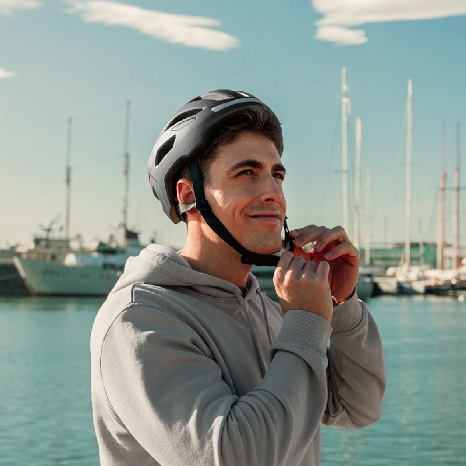 Un joven con sudadera gris ajusta su casco de bicicleta frente a un puerto y barcos.