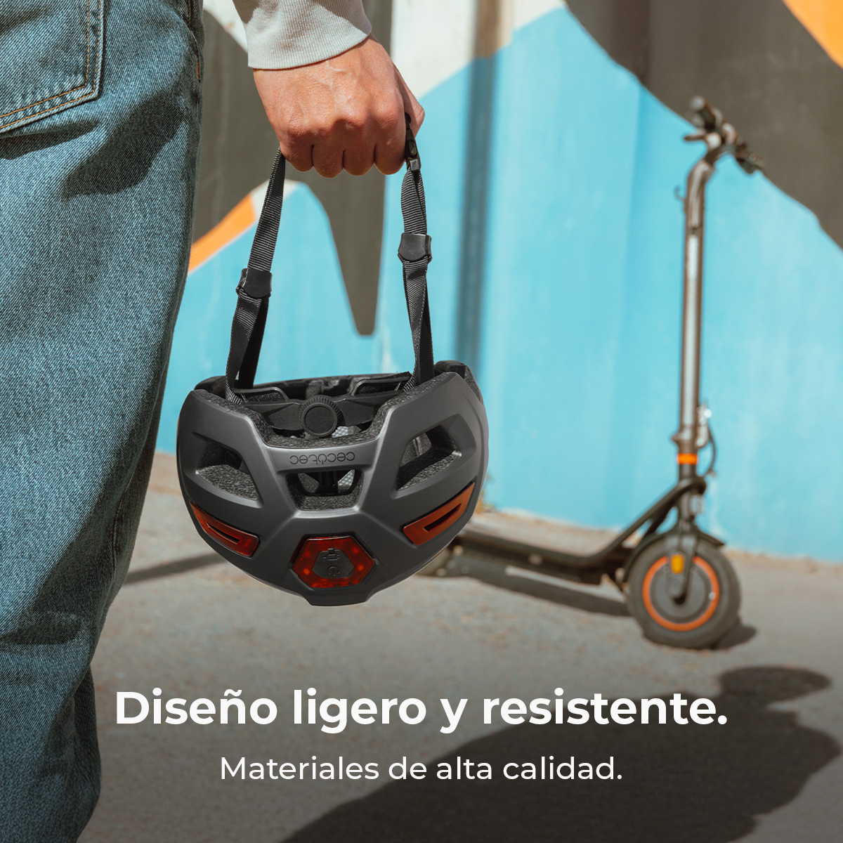 Persona con jeans sostiene un casco negro con luces integradas frente a un fondo azul y naranja con un patinete.