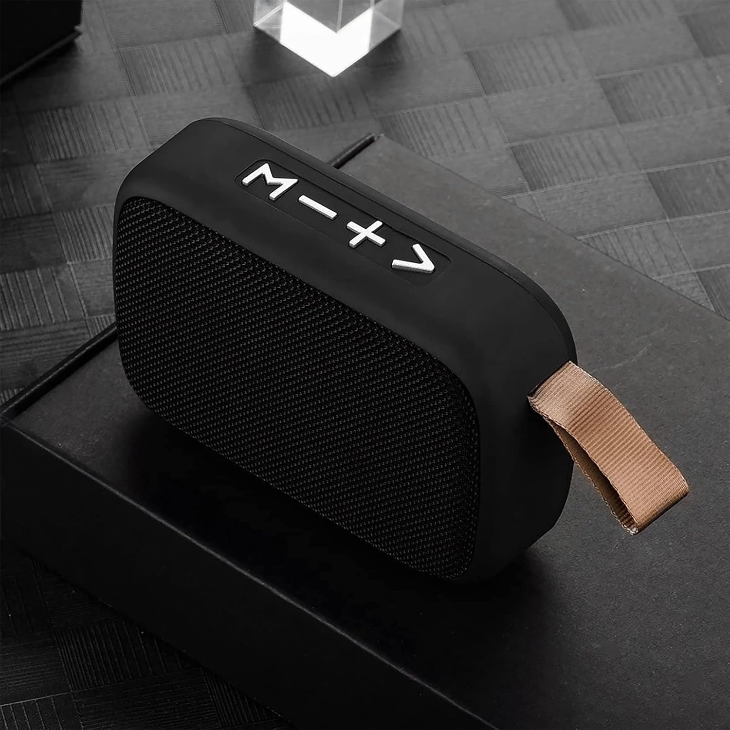 Ein schwarzer, tragbarer Bluetooth-Lautsprecher mit weißer Beschriftung und einer braunen Lasche auf einer dunklen Oberfläche.