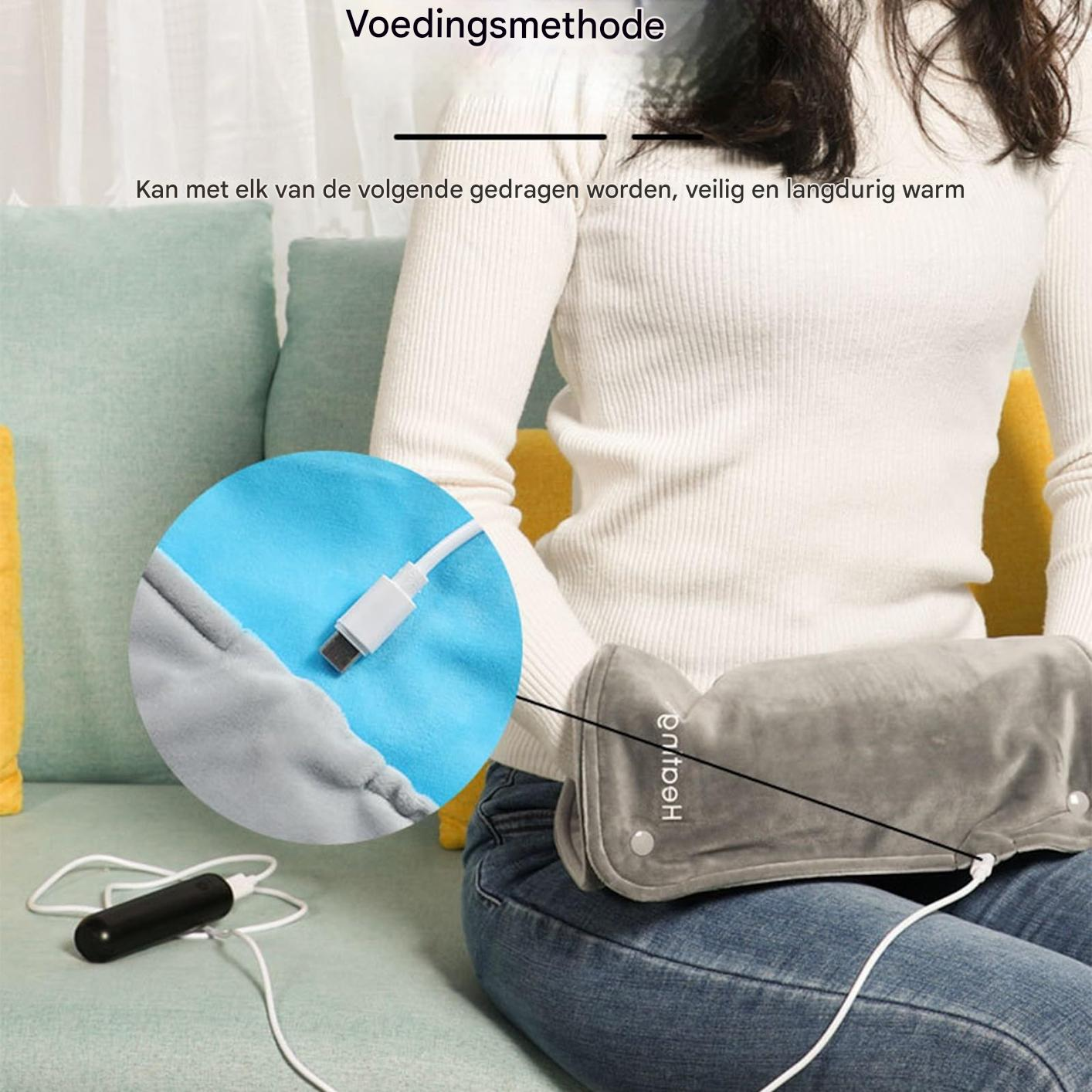 Eine Person auf einem grünen Sofa hält einen grauen, beheizbaren Handwärmer, der mit einer Powerbank verbunden ist.