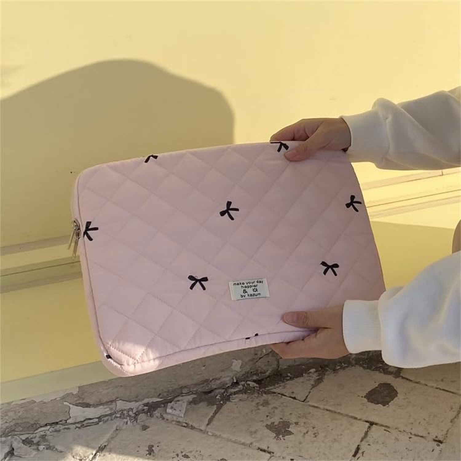 Eine Person hält eine hellrosa, gesteppte Laptoptasche mit kleinen schwarzen Schleifen vor einer Wand.