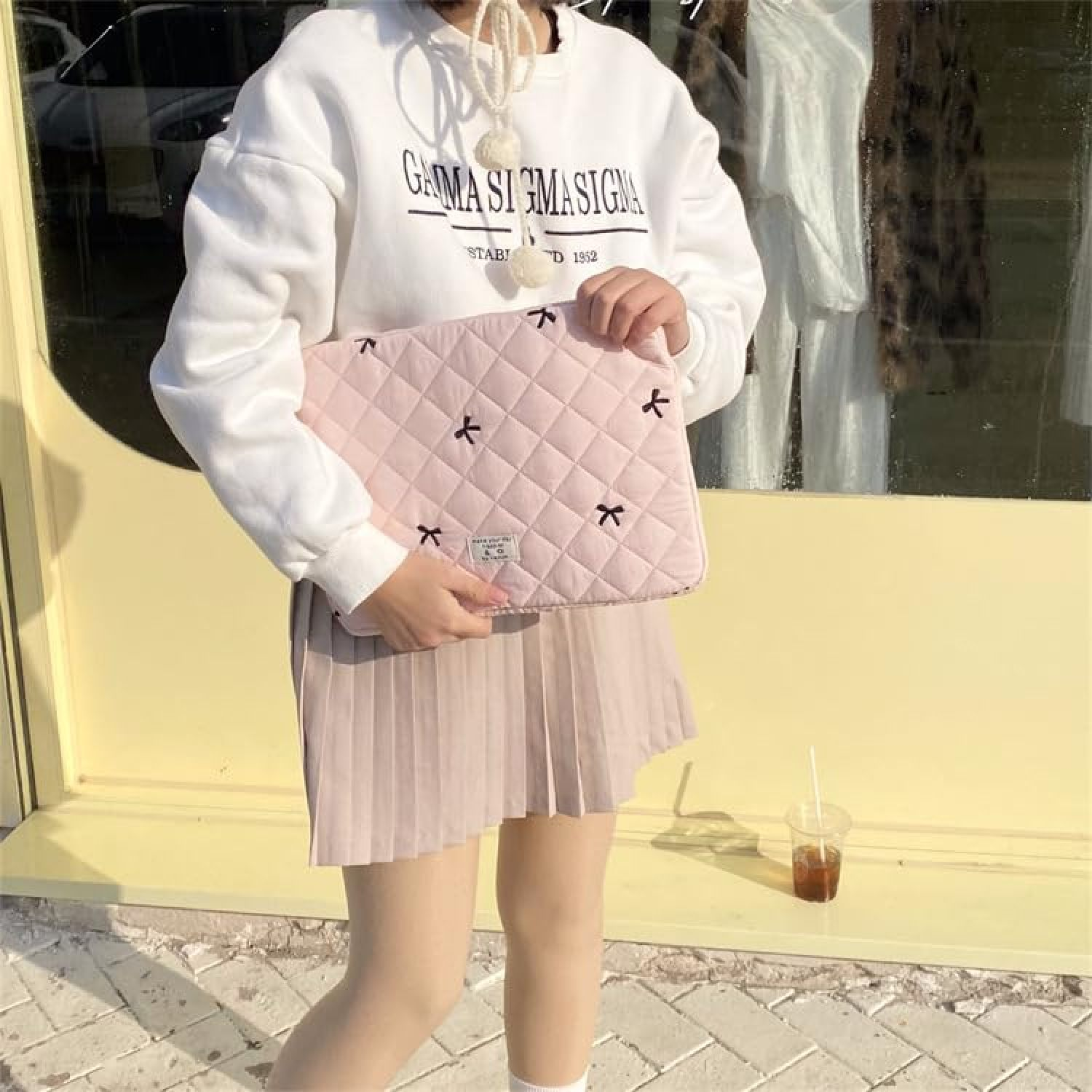 Eine Frau in weißem Pullover und beiger Faltenrock hält eine rosa gesteppte Laptoptasche mit schwarzen Schleifen.