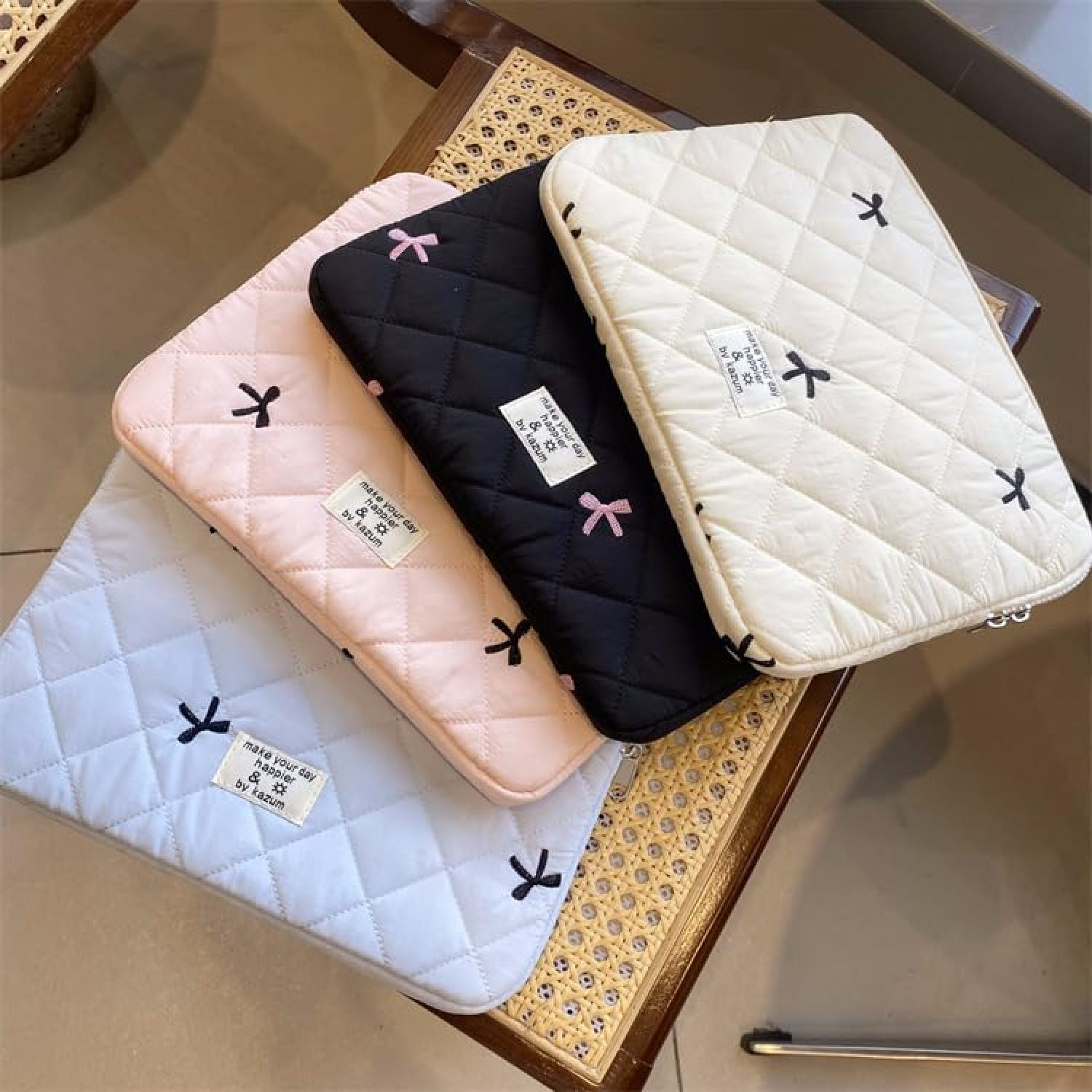 Vier gesteppte Laptoptaschen in Hellblau, Rosa, Schwarz und Weiß auf einem geflochtenen Tisch.