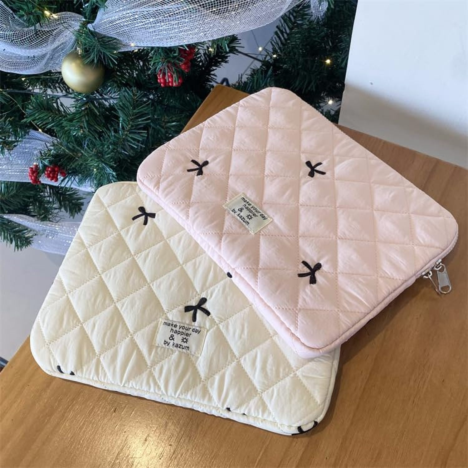 Zwei gesteppte Laptop-Hüllen in Rosa und Creme mit kleinen Schleifen und Etikett auf Holz-Tisch.