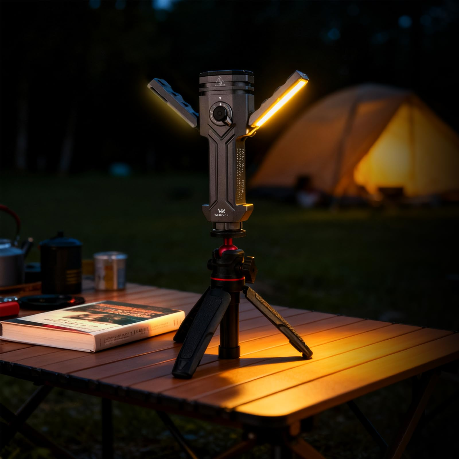 Eine graue LED-Campinglampe auf einem Mini-Stativ auf einem Holztisch, im Hintergrund ein beleuchtetes Zelt bei Nacht.