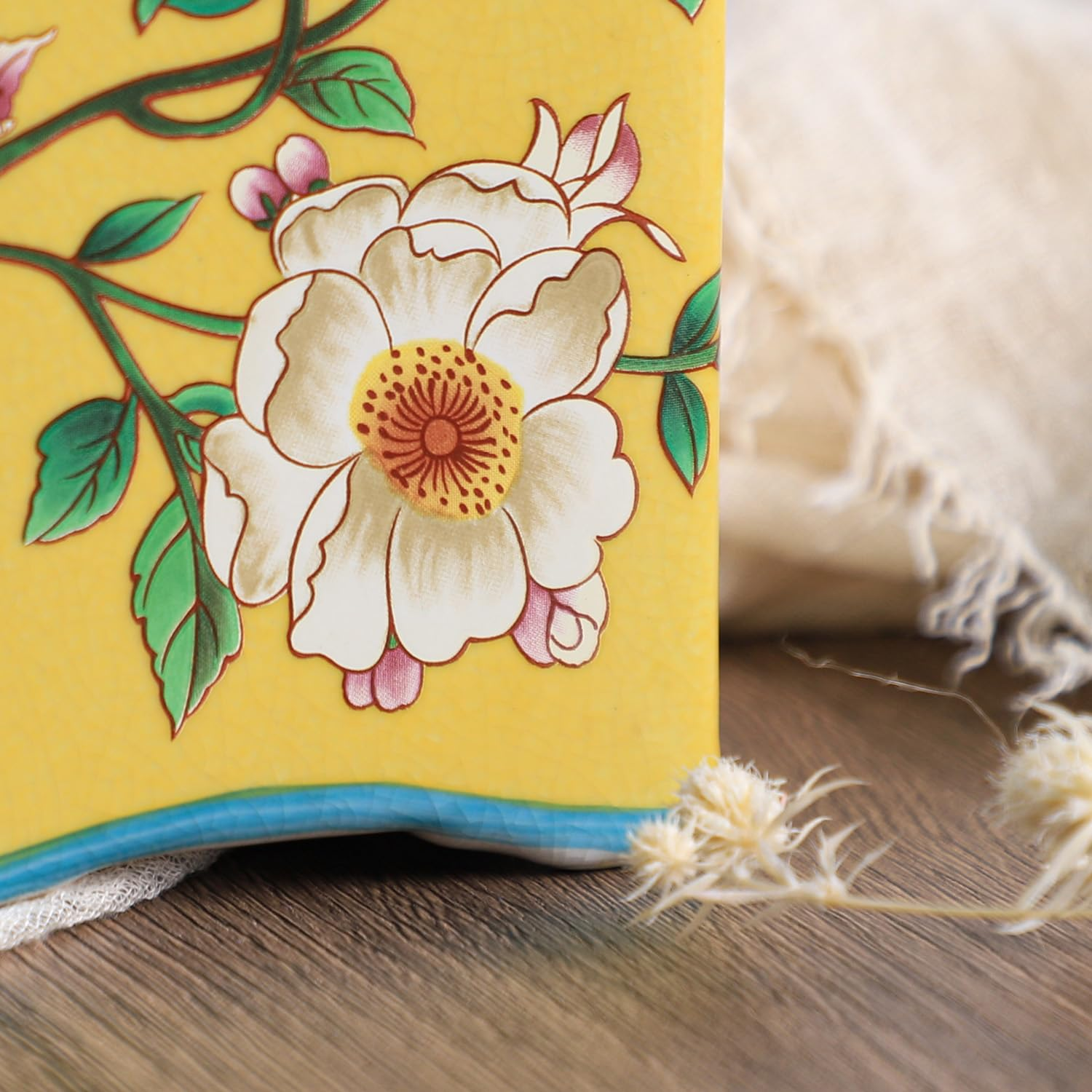 Nahaufnahme einer gelben Keramikvase mit bemalten weißen Blumen, grünen Blättern und roten Details vor Textil.