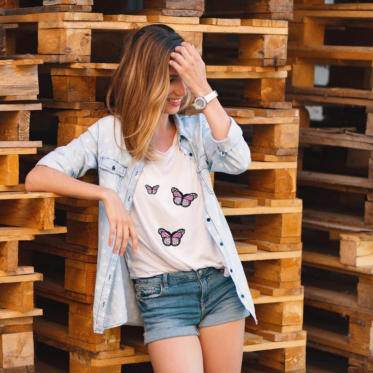 Eine Frau stützt sich auf Holzpaletten und trägt ein Schmetterlings-T-Shirt, ein hellblaues Hemd und Jeansshorts.