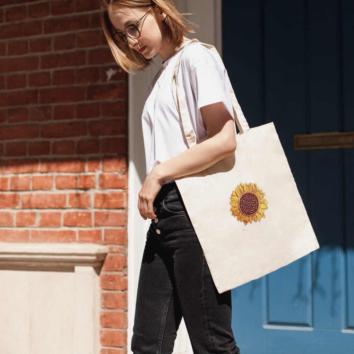 Eine Frau in weißem Shirt und Jeans trägt eine helle Stofftasche mit Sonnenblumen-Motiv.