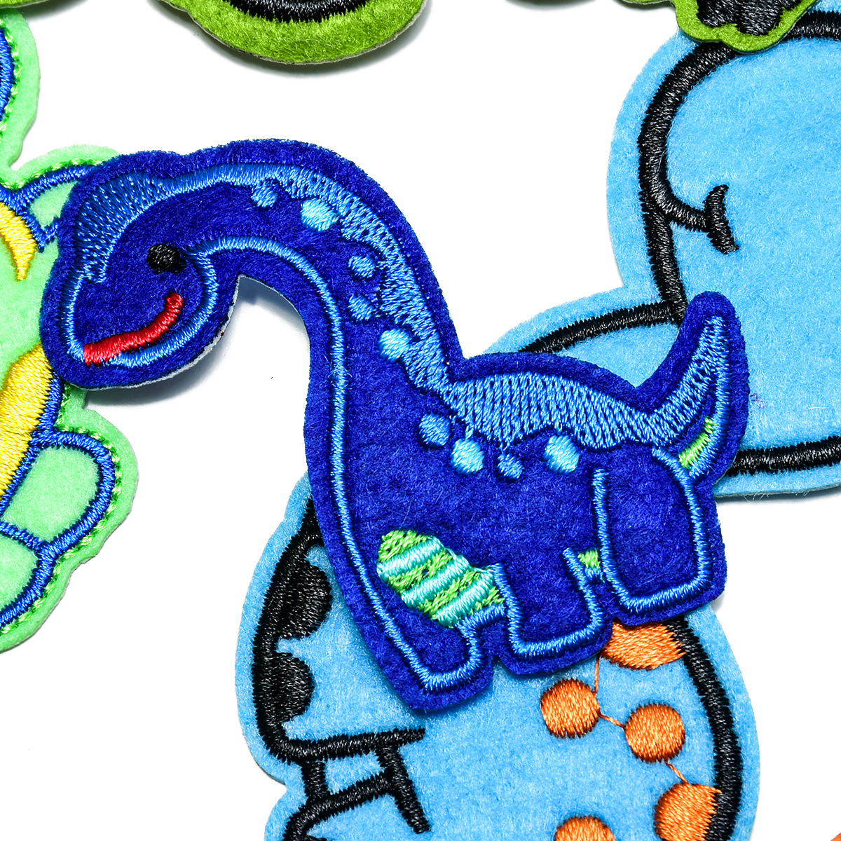 Ein blauer, bestickter Dinosaurier-Aufnäher auf weißem Hintergrund, umgeben von anderen bunten Filzaufnähern.