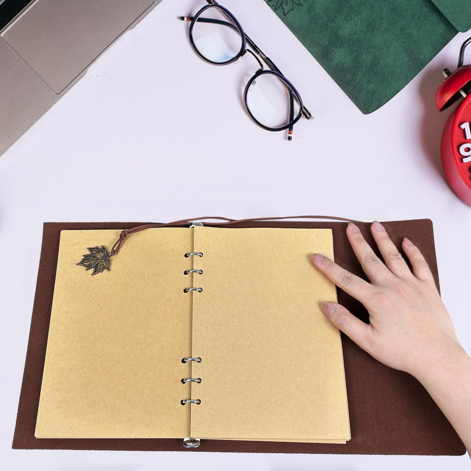 Hand berührt ein offenes Notizbuch mit braunem Einband auf hellem Tisch mit Brille und Wecker.