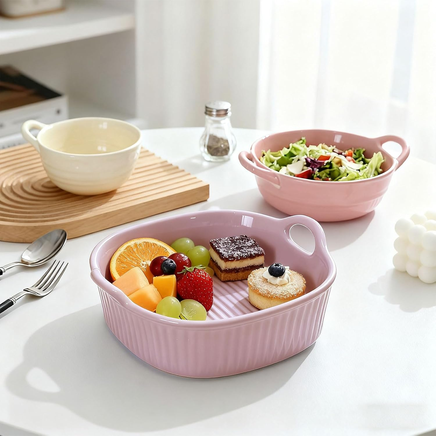 Eine rosa Schale mit Obst und Kuchen, dazu eine Salatschüssel und ein Becher auf weißem Tisch.
