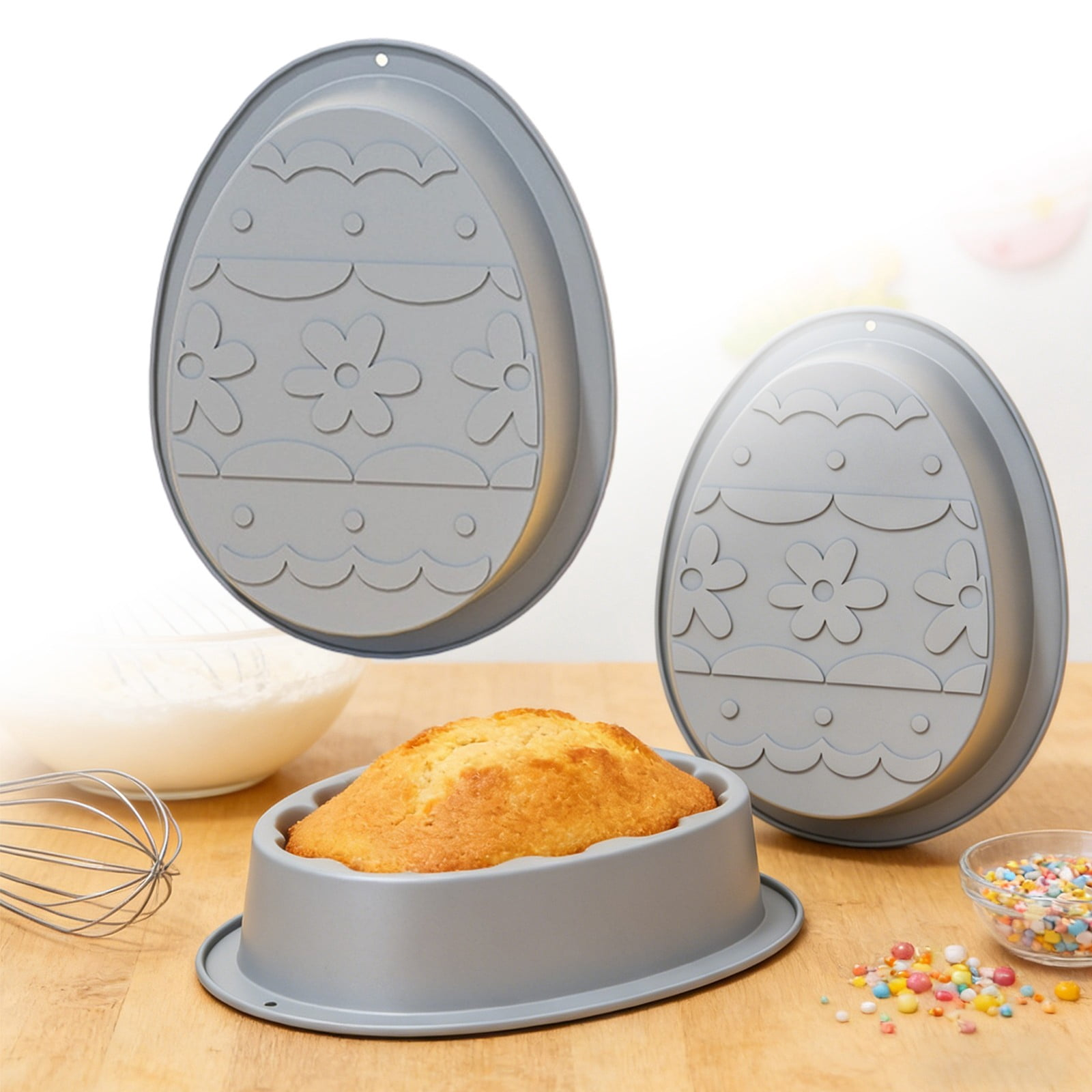 Graue Eier-Backformen auf Holztisch, eine mit gebackenem Kuchen, Schneebesen, Schüssel und bunten Streuseln.