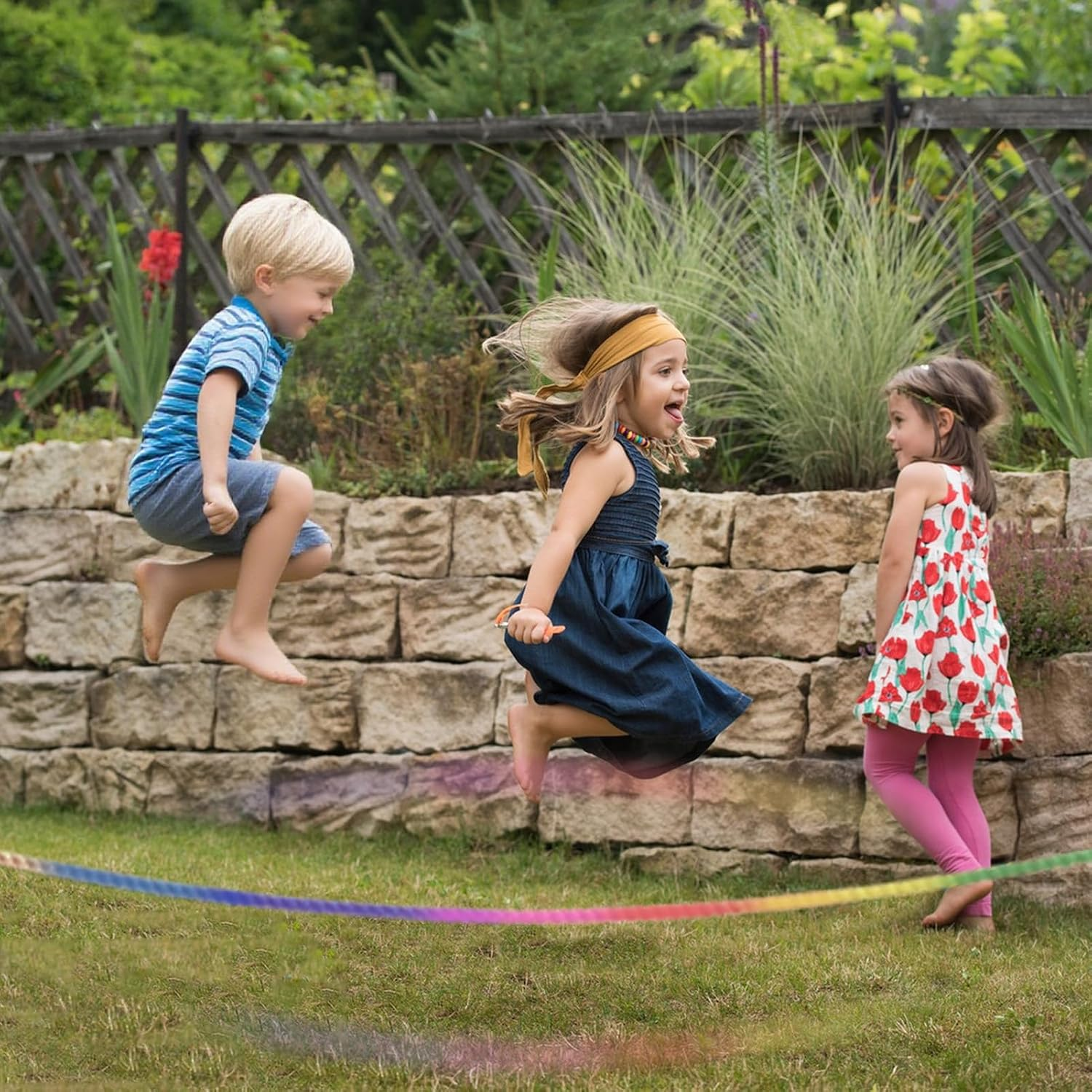 Drei Kinder springen in einem Garten über ein buntes Seil vor einer Steinmauer.