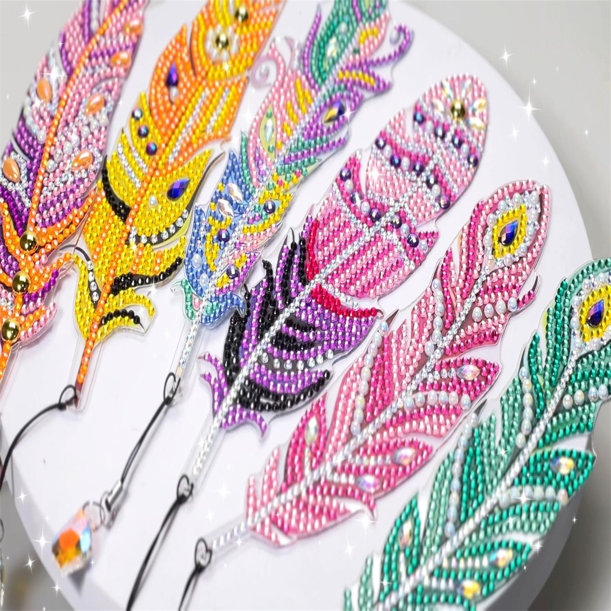 Sechs bunte, glitzernde Diamantmalerei-Lesezeichen in Federform liegen auf einer weißen Fläche.