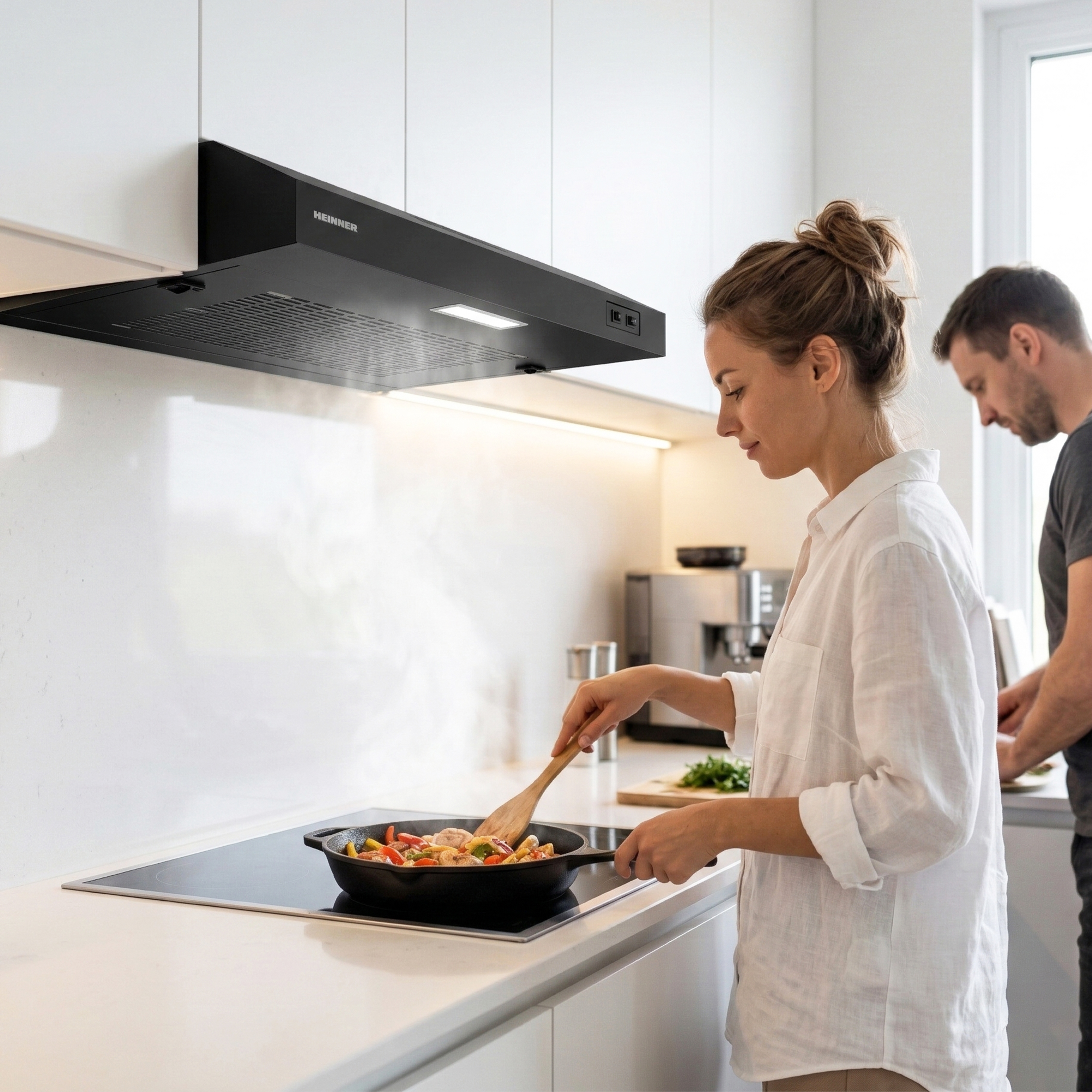 Une femme cuisine des légumes dans une poêle dans une cuisine moderne pendant qu'un homme se tient en arrière-plan.