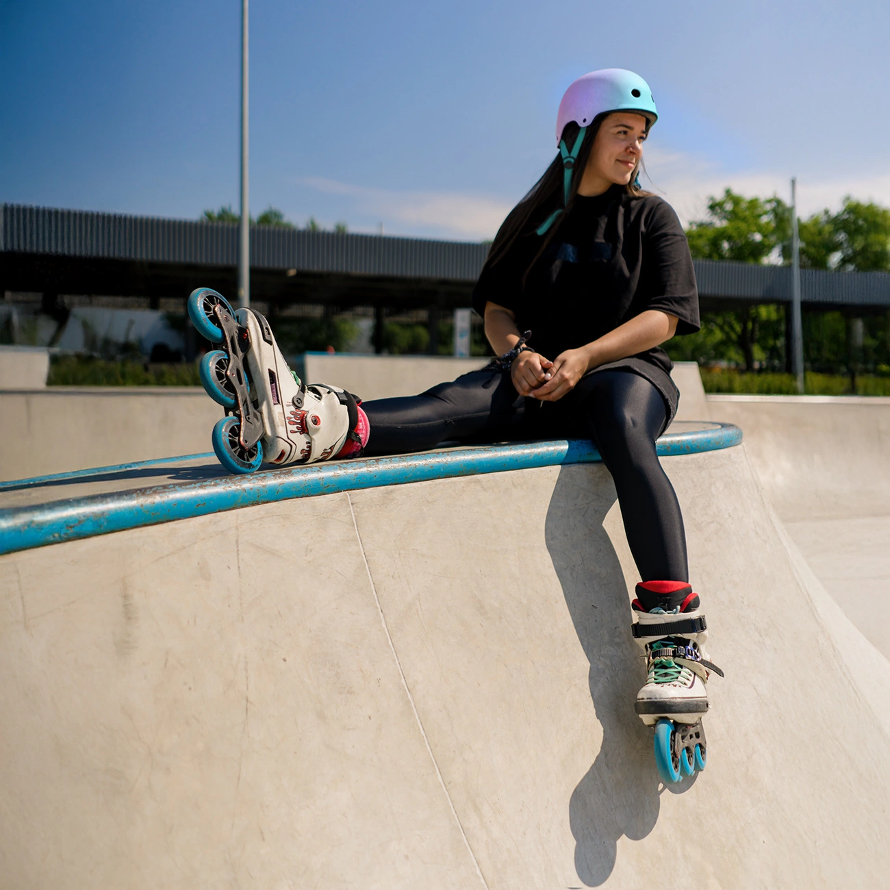 Una giovane donna con casco e pattini in linea siede su una rampa di uno skatepark in una giornata di sole.