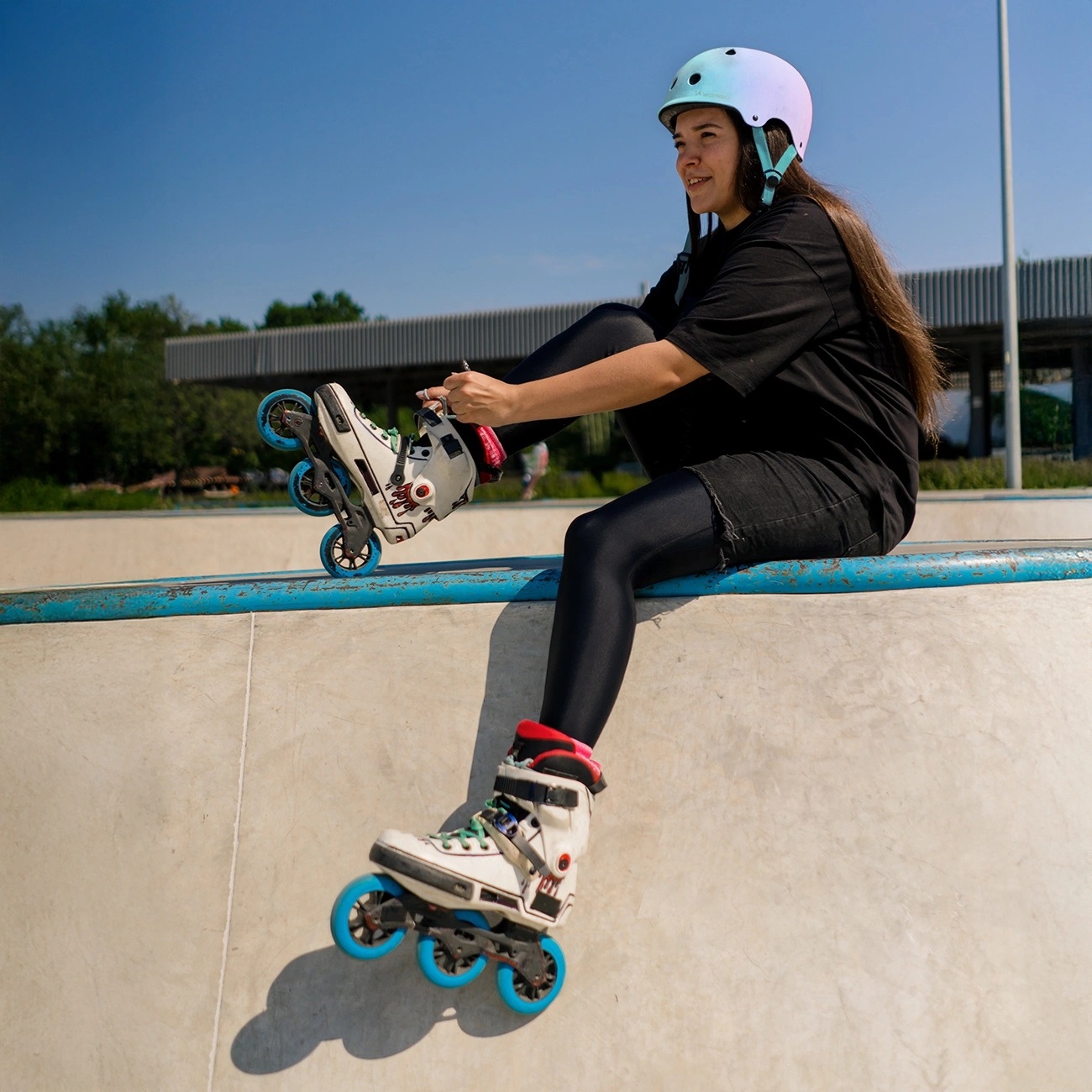 Una giovane donna con il casco siede sul muretto di uno skatepark mentre indossa pattini in linea bianchi con ruote blu.