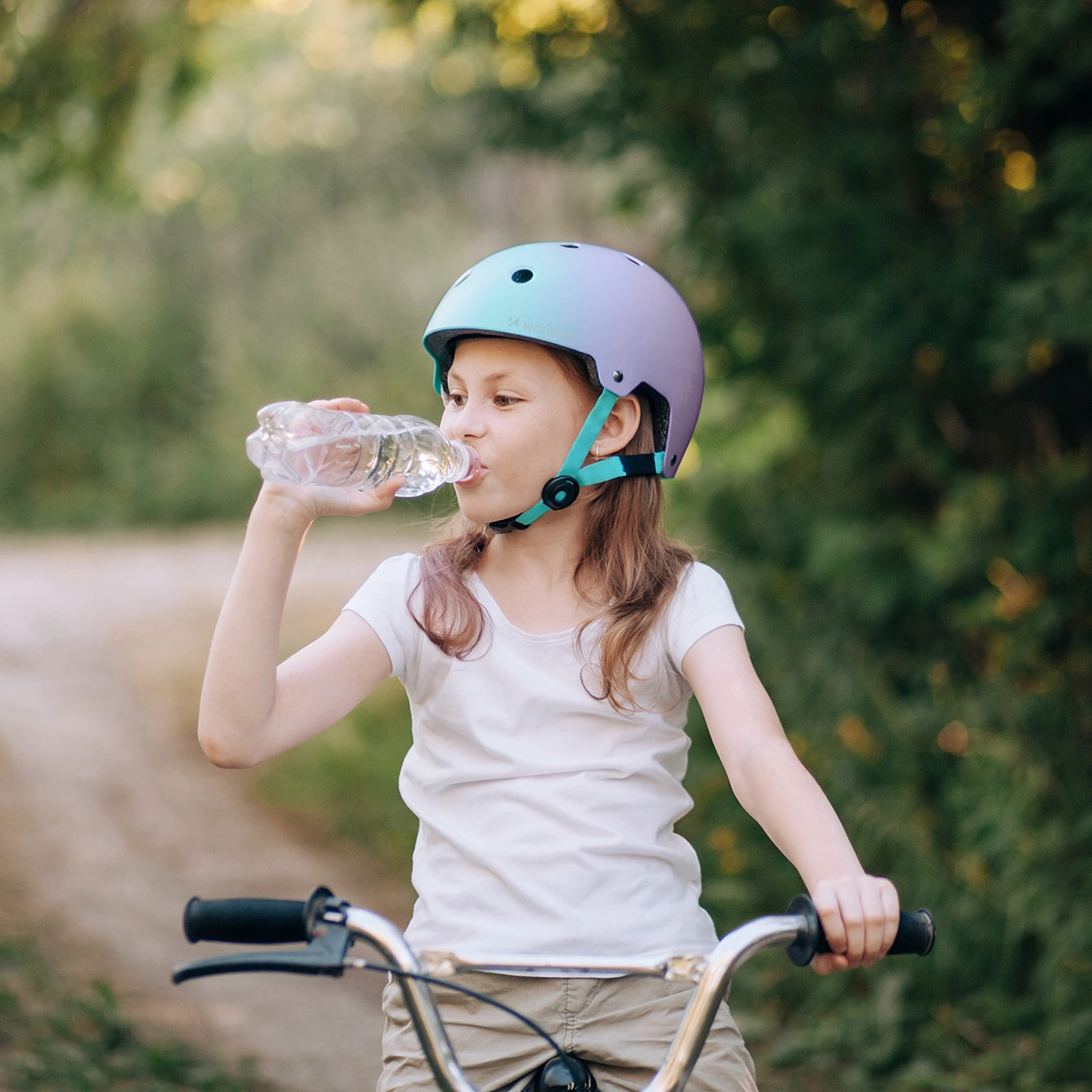 Una bambina con casco blu e viola beve acqua da una bottiglia mentre va in bicicletta all'aperto.