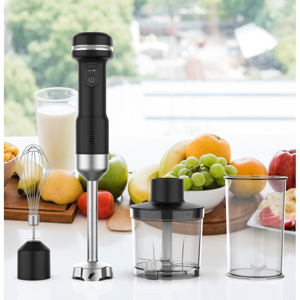 Czarny blender ręczny z akcesoriami, takimi jak trzepaczka, rozdrabniacz i pojemnik, w otoczeniu świeżych owoców.