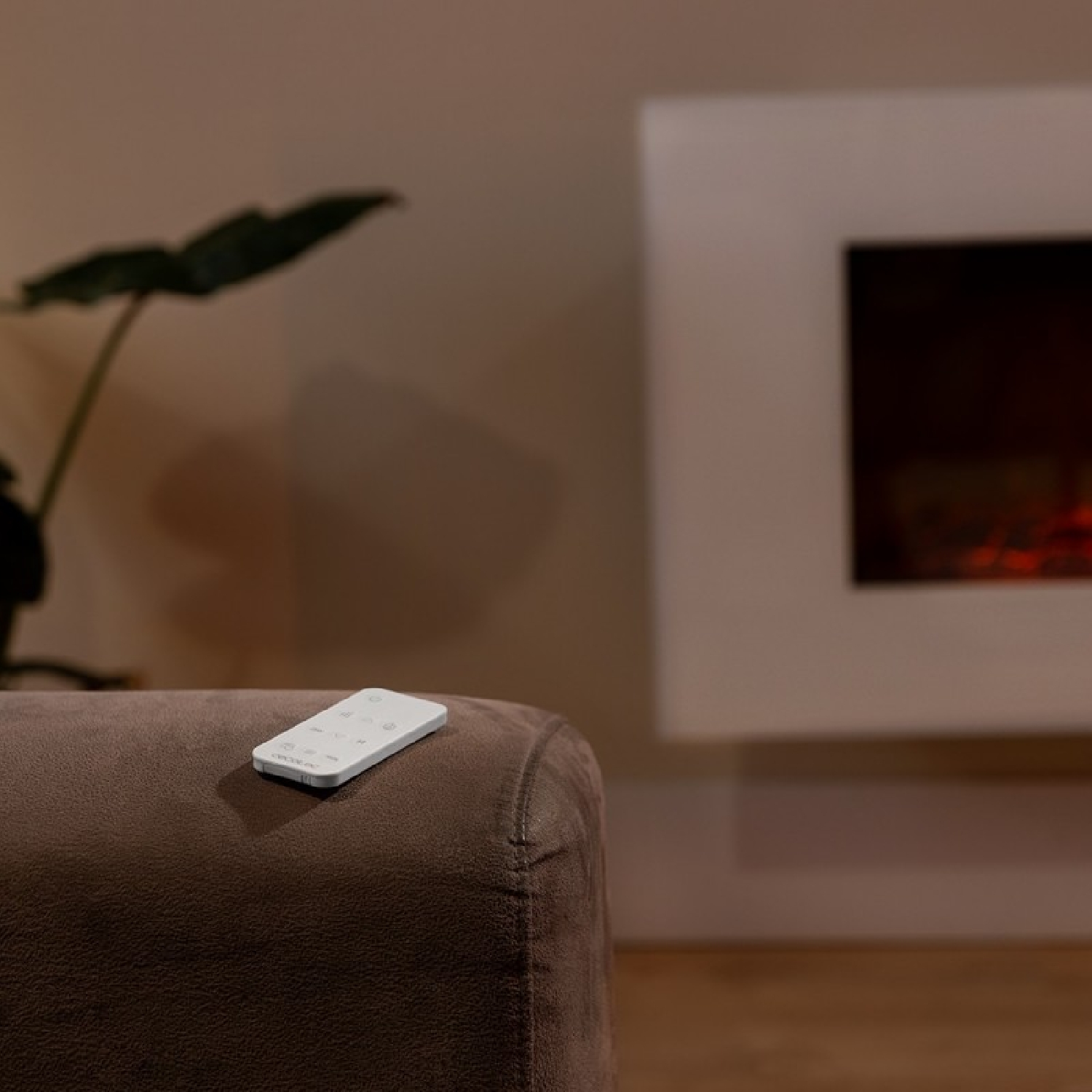 Un mando a distancia blanco sobre un sofá marrón, con una chimenea y una planta al fondo.