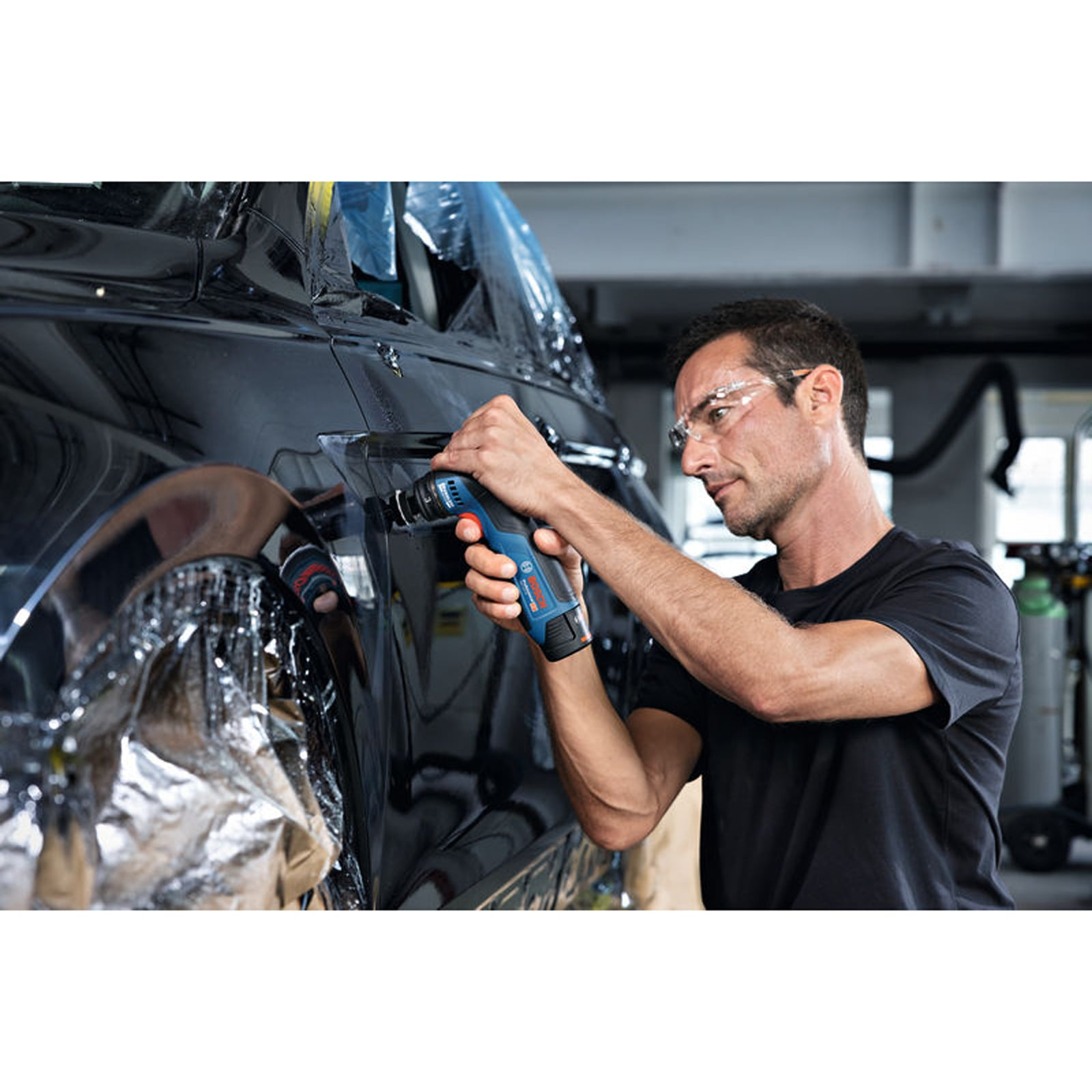 Ein Handwerker mit Schutzbrille schneidet mit einer Akku-Multi-Säge ein schwarzes Autoteil zu.
