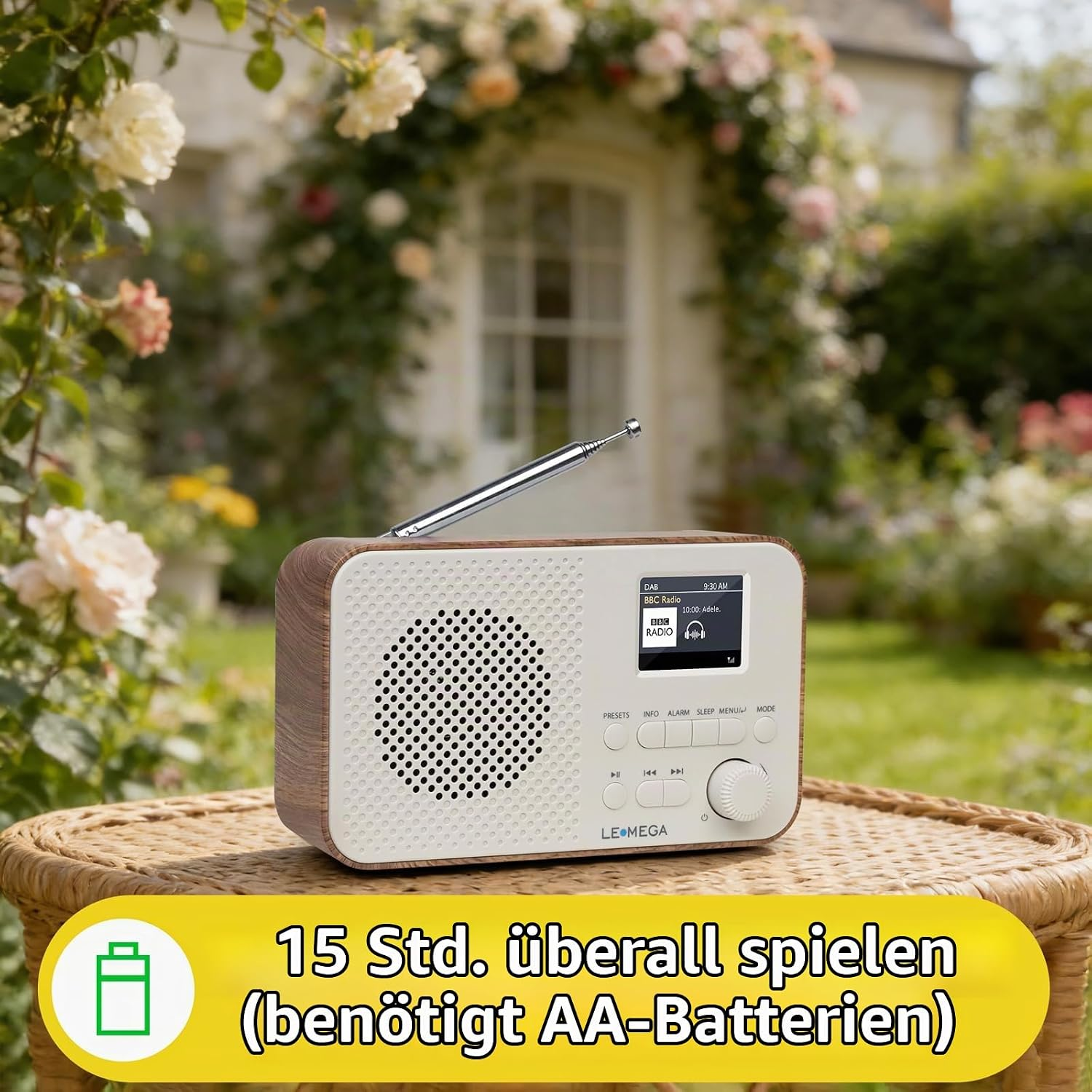 Ein weißes Retro-Radio mit Holzrahmen auf einem Rattantisch im Garten vor einem blühenden Rosenbogen.