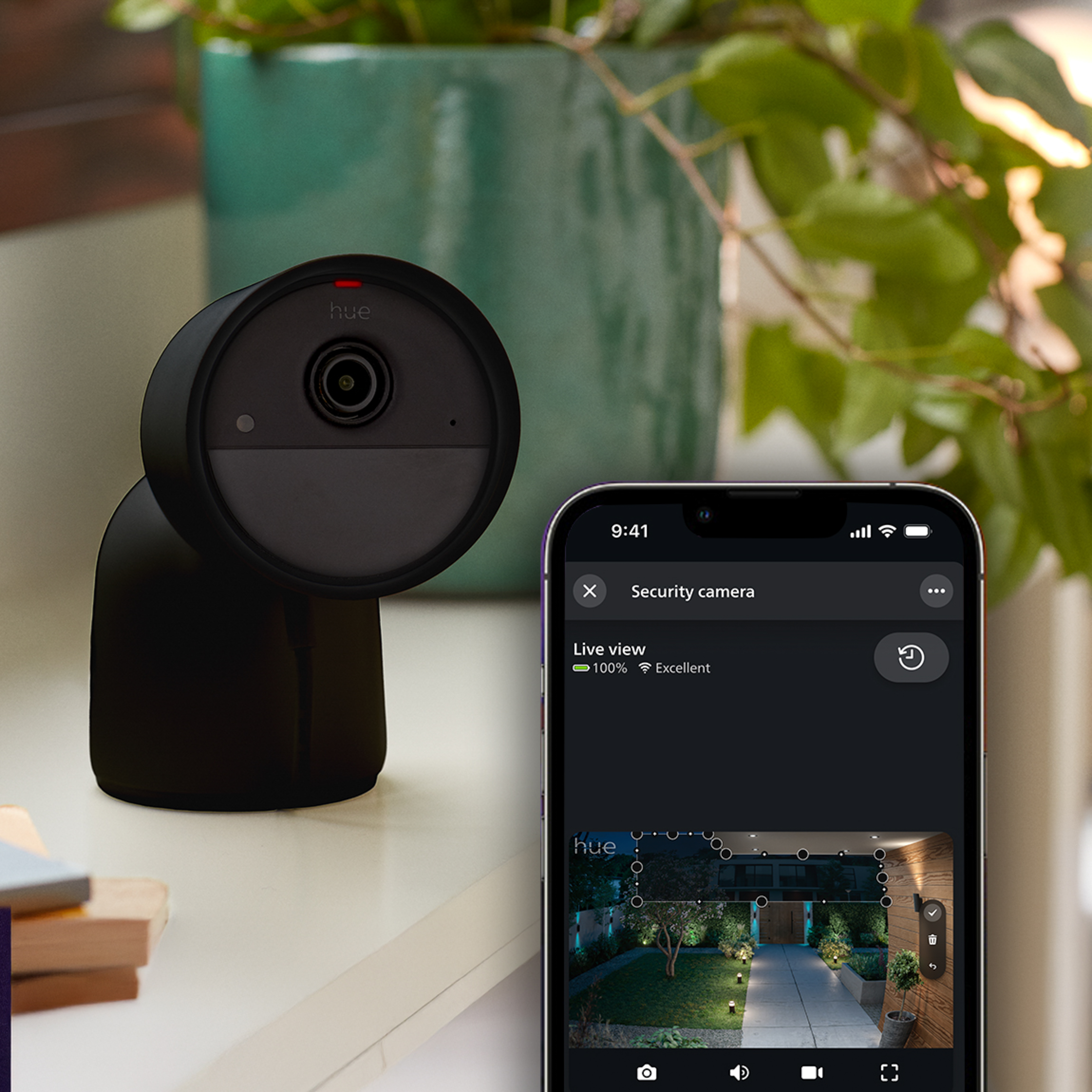 Schwarze Hue Sicherheitskamera auf einer Oberfläche neben einem Smartphone, das eine Live-Ansicht zeigt.