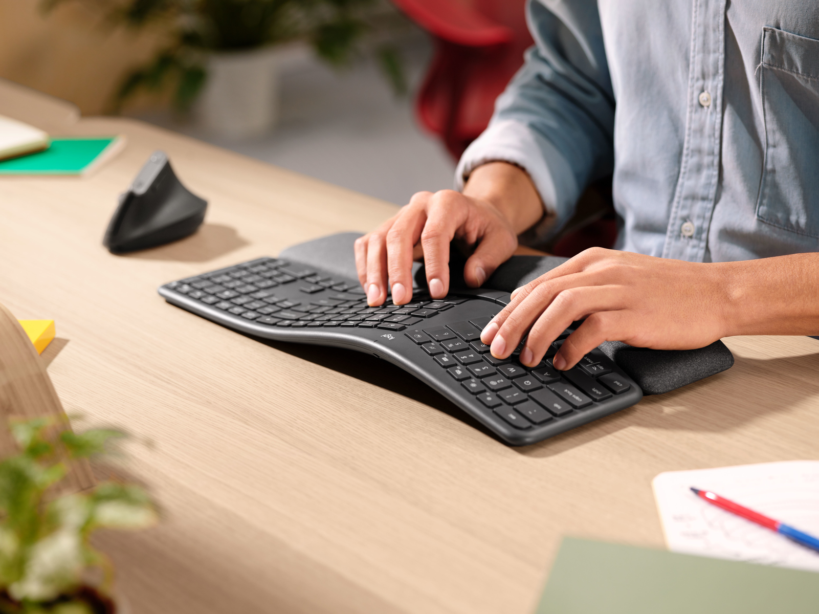 Person tippt auf einer ergonomischen, geschwungenen schwarzen Tastatur mit Handballenauflage an einem Holztisch.