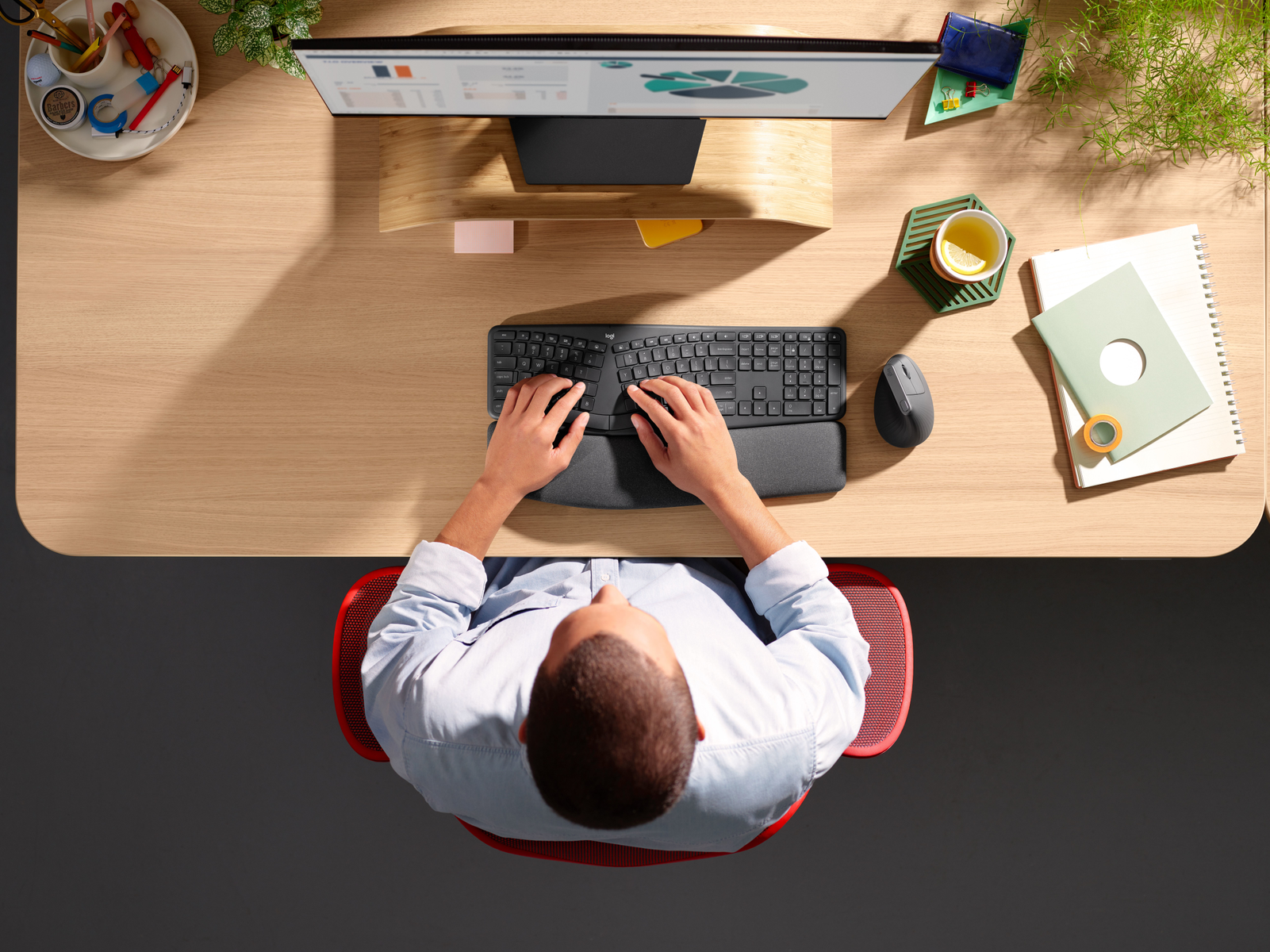 Draufsicht auf einen Schreibtisch mit Monitor, einer Person, die an einer ergonomischen Tastatur tippt, und Bürobedarf.