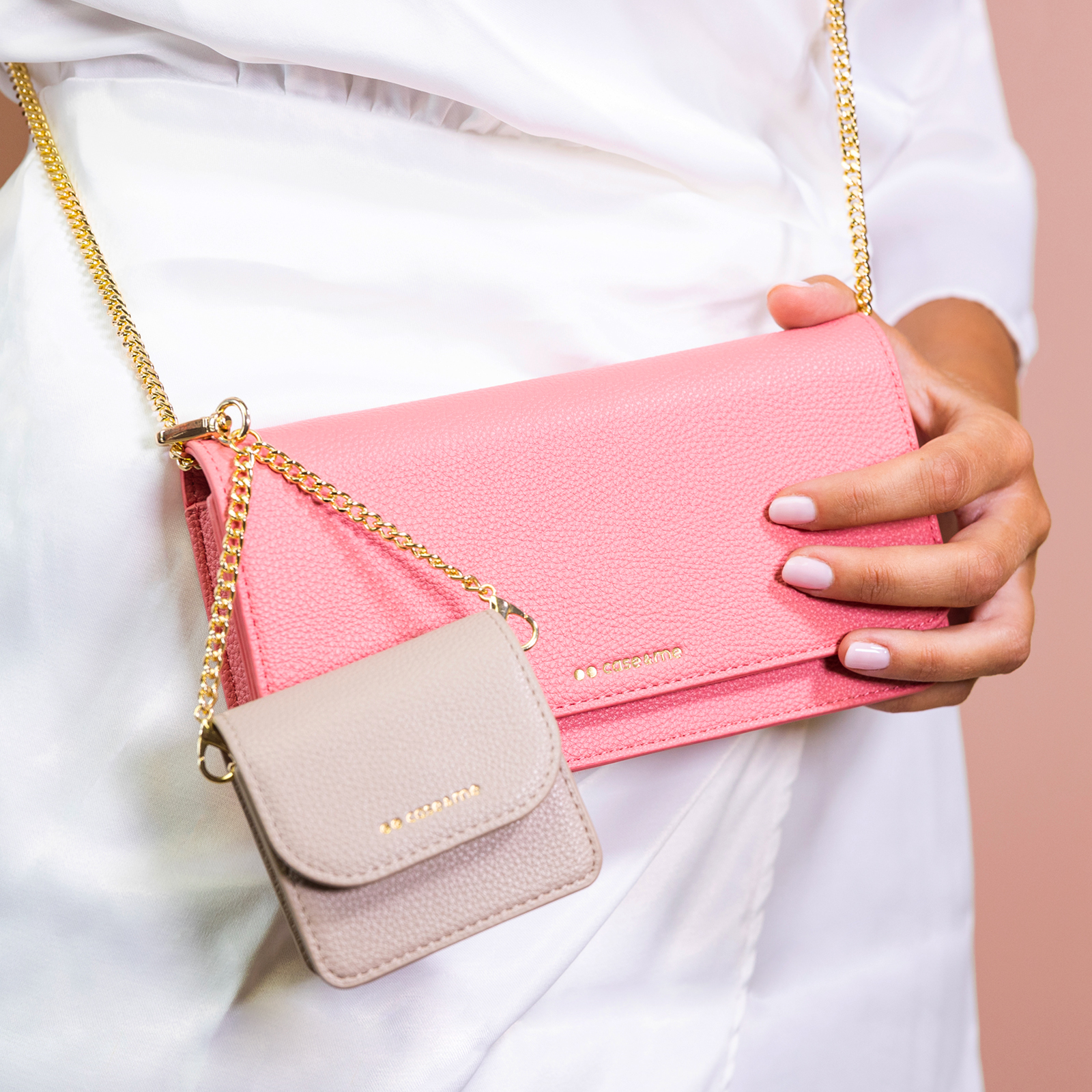 Una persona de blanco sostiene un bolso cruzado de cuero rosa con cadena dorada y un pequeño monedero beige.