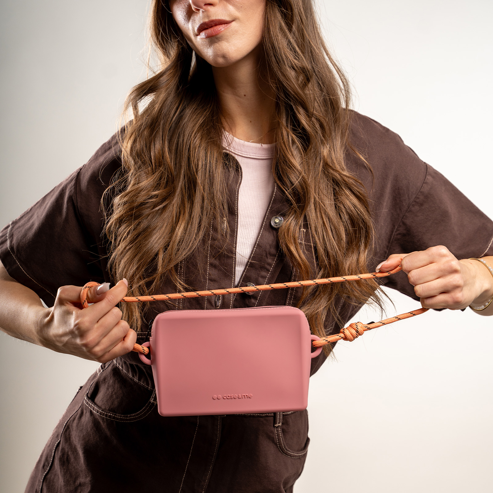 Una mujer con ropa marrón sostiene un pequeño bolso cruzado rosa con correa de cuerda naranja.