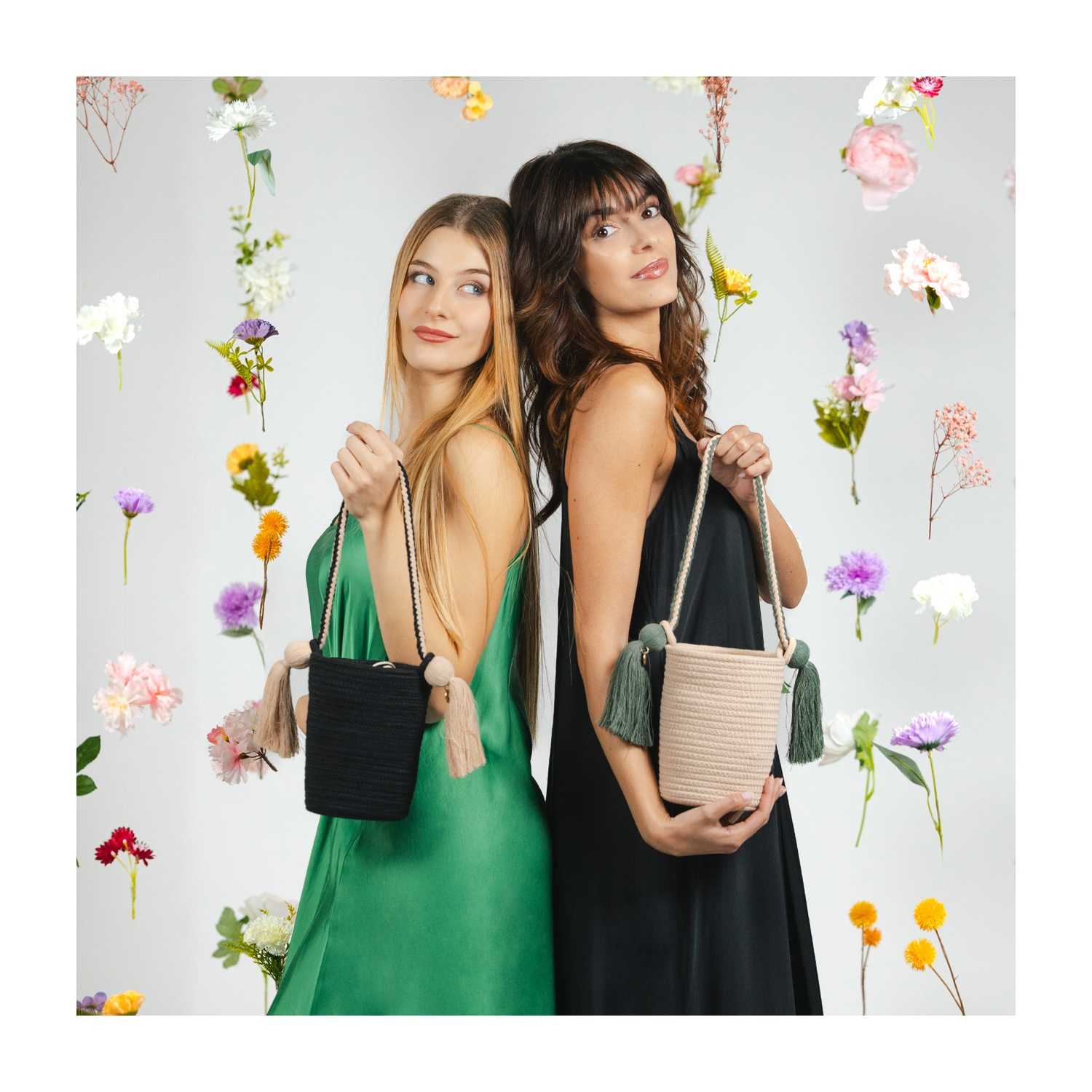 Dos mujeres con vestidos verde y negro muestran bolsos ante flores flotantes.