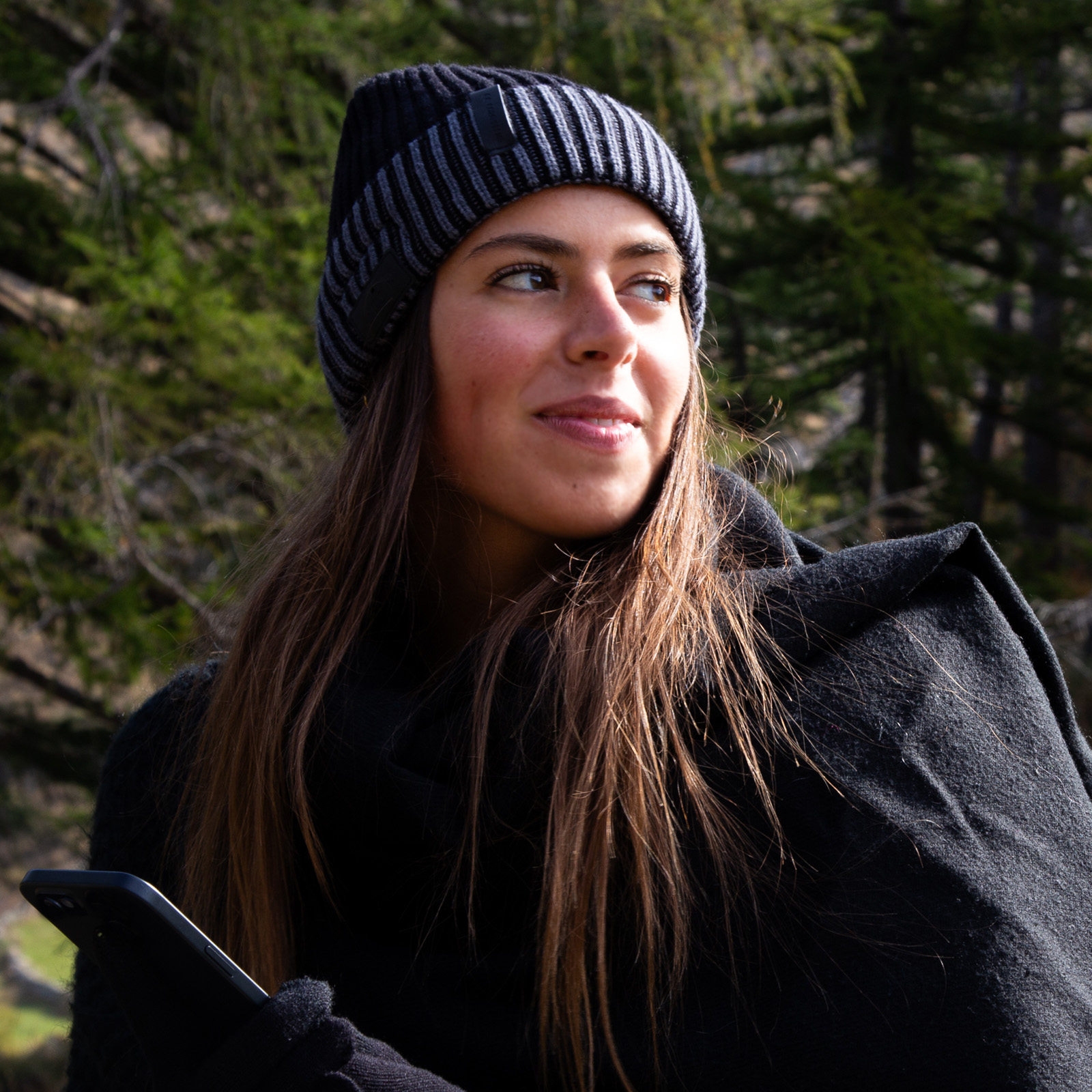 Una donna con berretto e cappotto nero tiene in mano un telefono all'aperto con alberi sfocati.