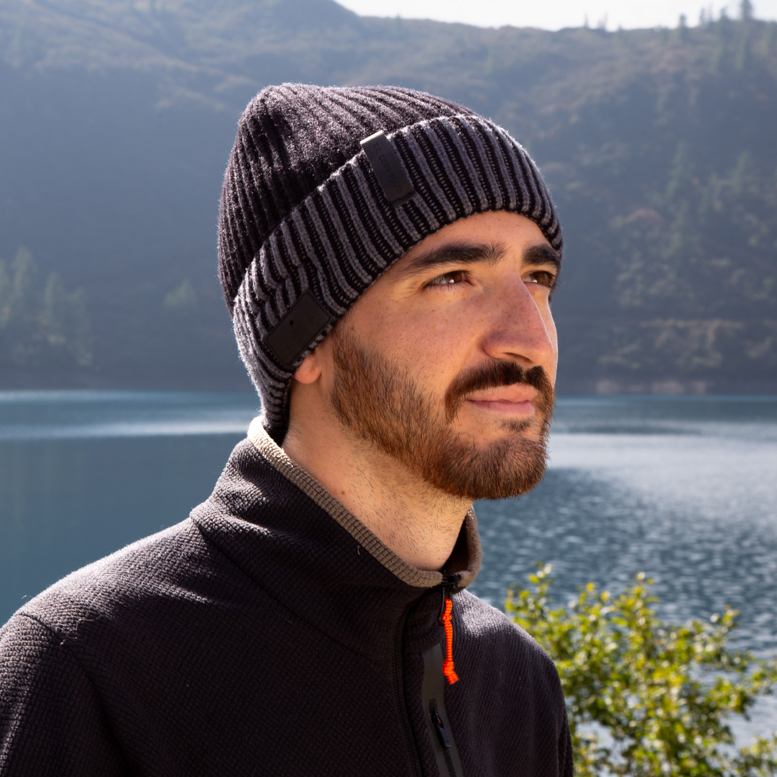 Un uomo con la barba indossa un berretto nero davanti a un lago e montagne boscose.