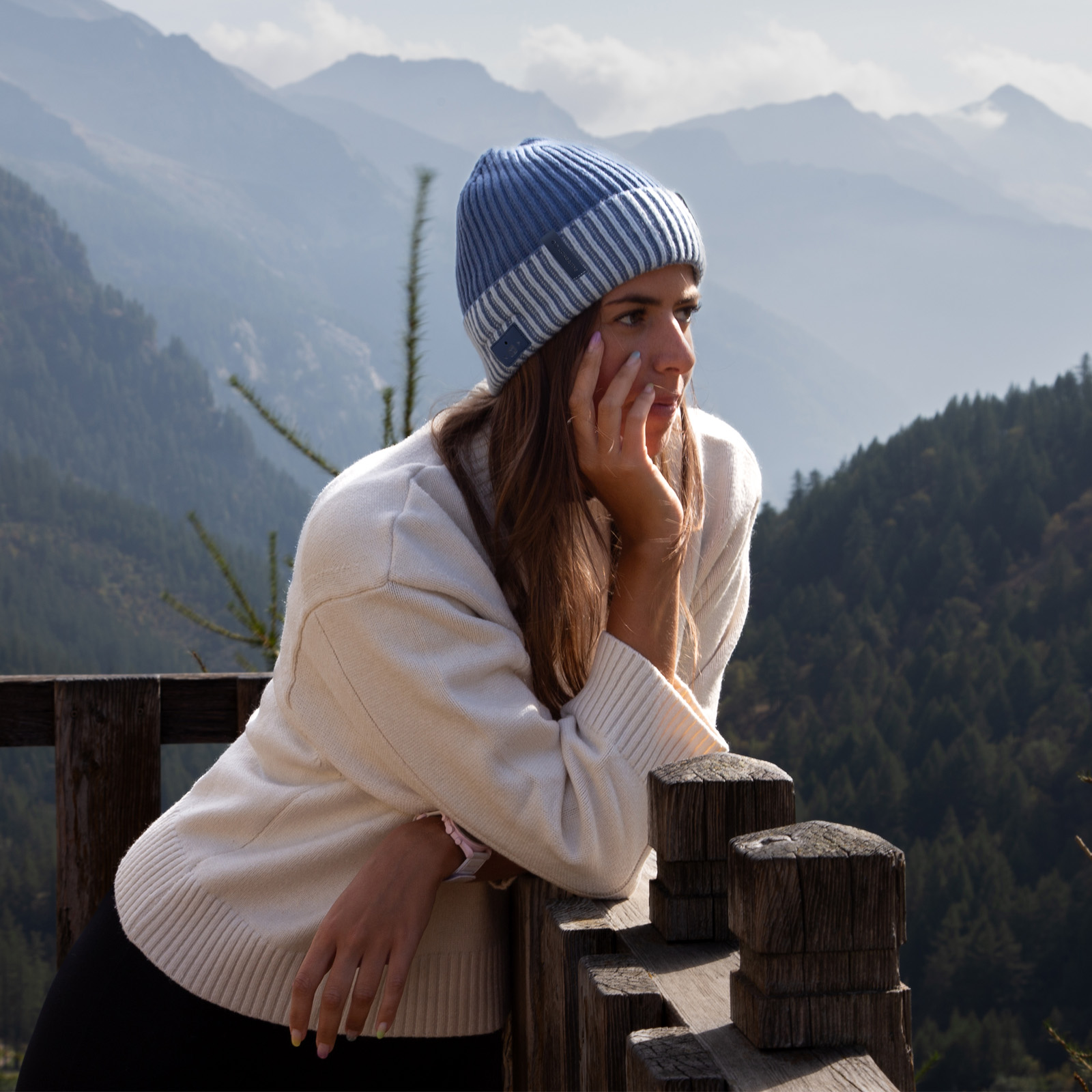 Una giovane donna con berretto blu e maglione bianco si appoggia a una ringhiera di legno tra i monti.